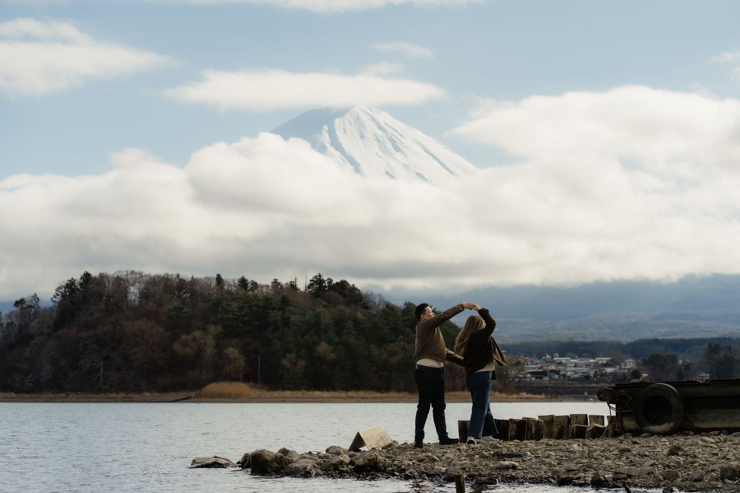 Two people stand by a rocky lake shore with snow-capped Mount Fuji rising behind clouds in Japan.