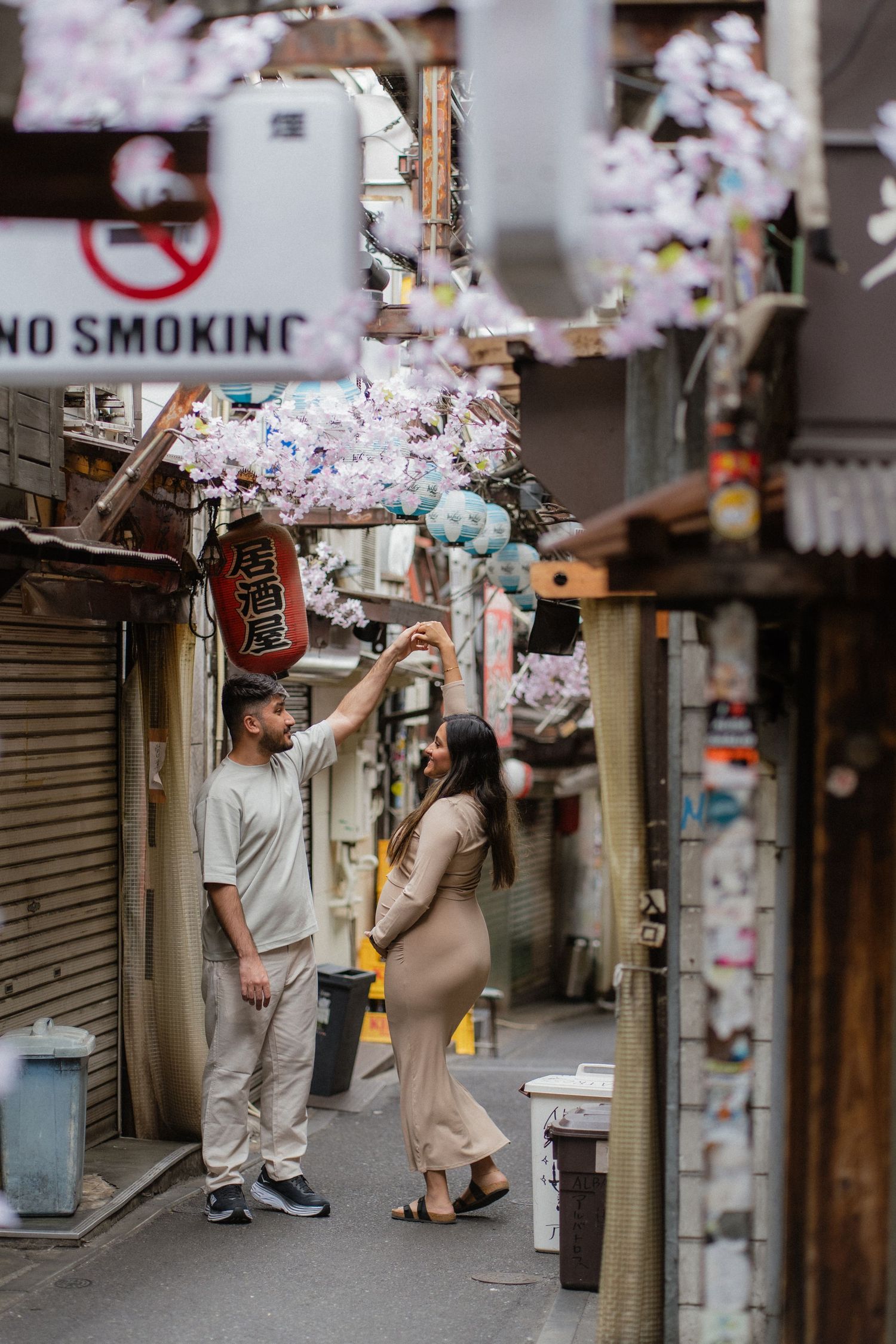 Couple touching cherry blossoms in a narrow Japanese alley with a no smoking sign overhead.