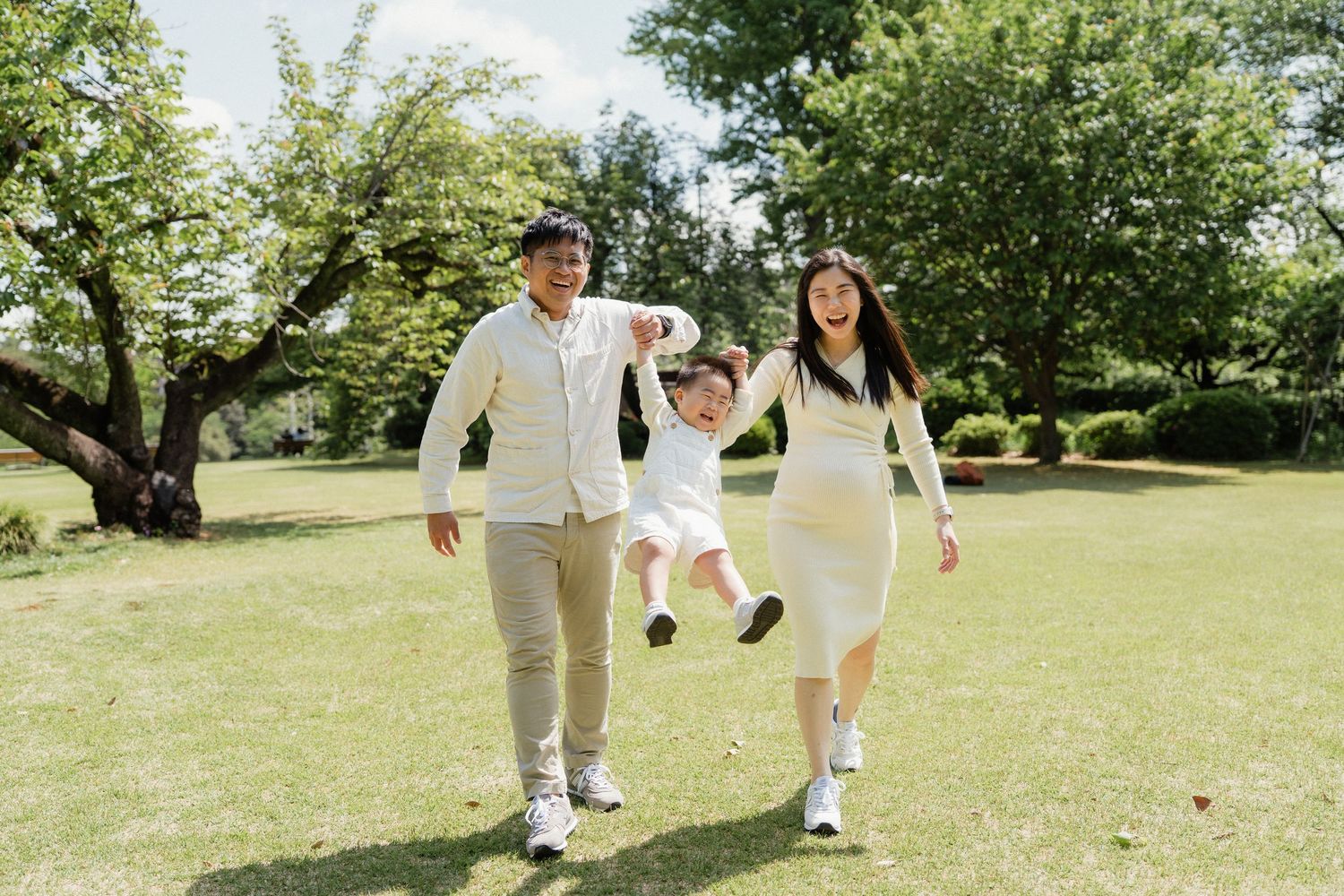 A joyful Asian family swinging their child between them while walking in a lush green park on a sunny day.