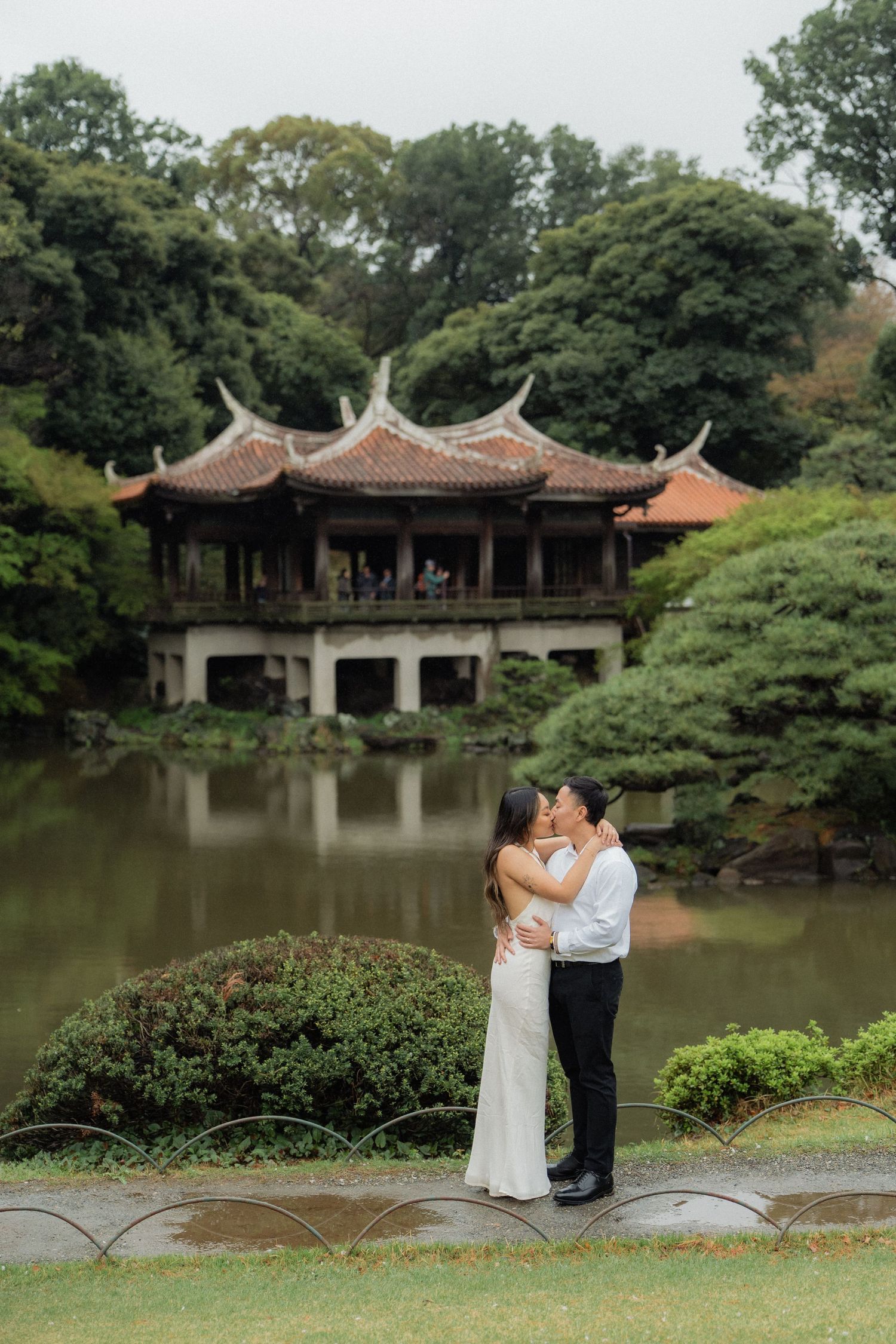 Couple sharing a kiss in white wedding attire before a serene Japanese garden pond with a traditional pagoda backdrop.