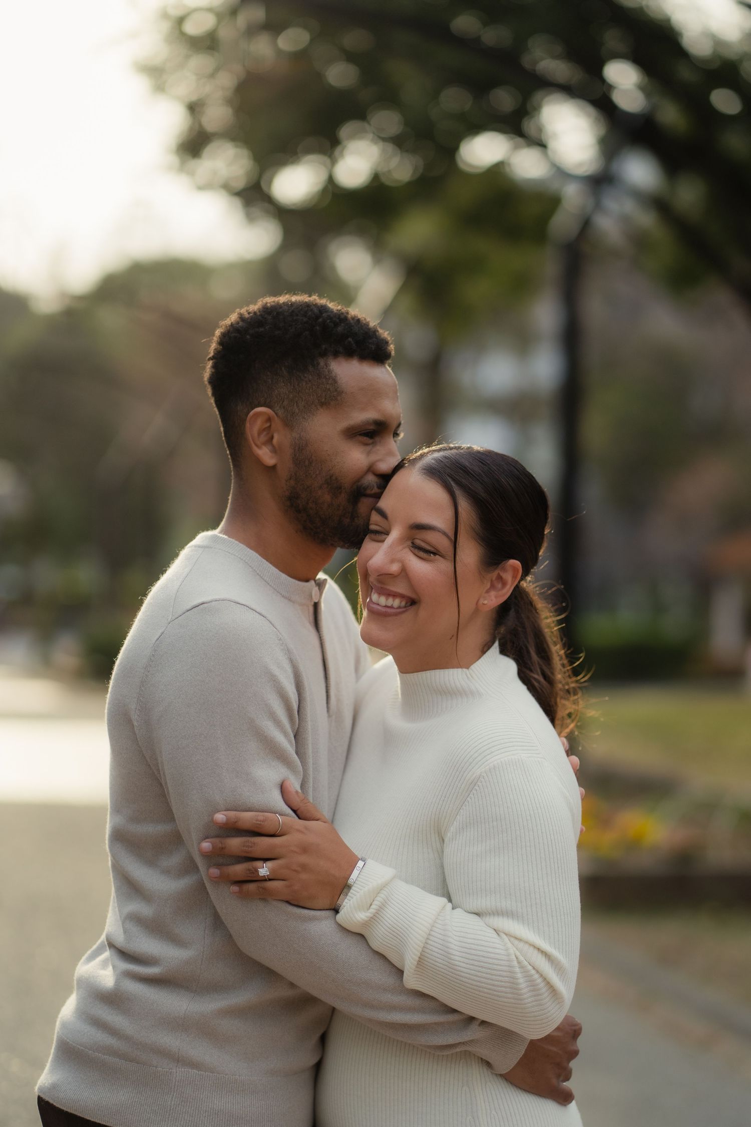 Happy couple embracing outdoors in a park, both wearing white sweaters, woman smiling joyfully.