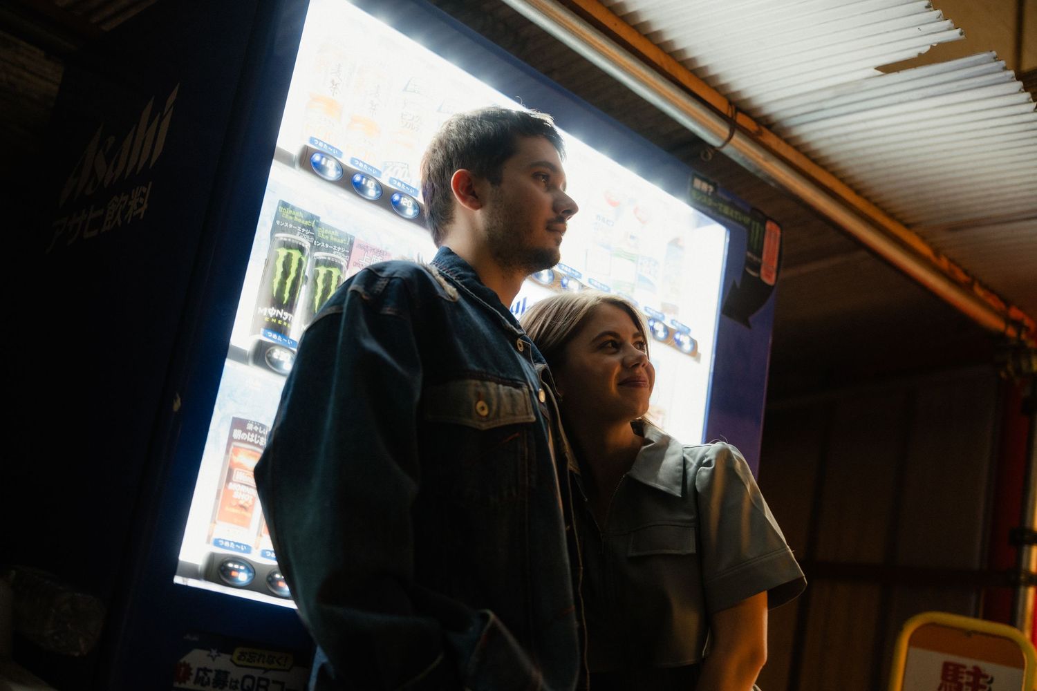 A young couple stands together in a dimly lit arcade, illuminated by the glow of a gaming screen.