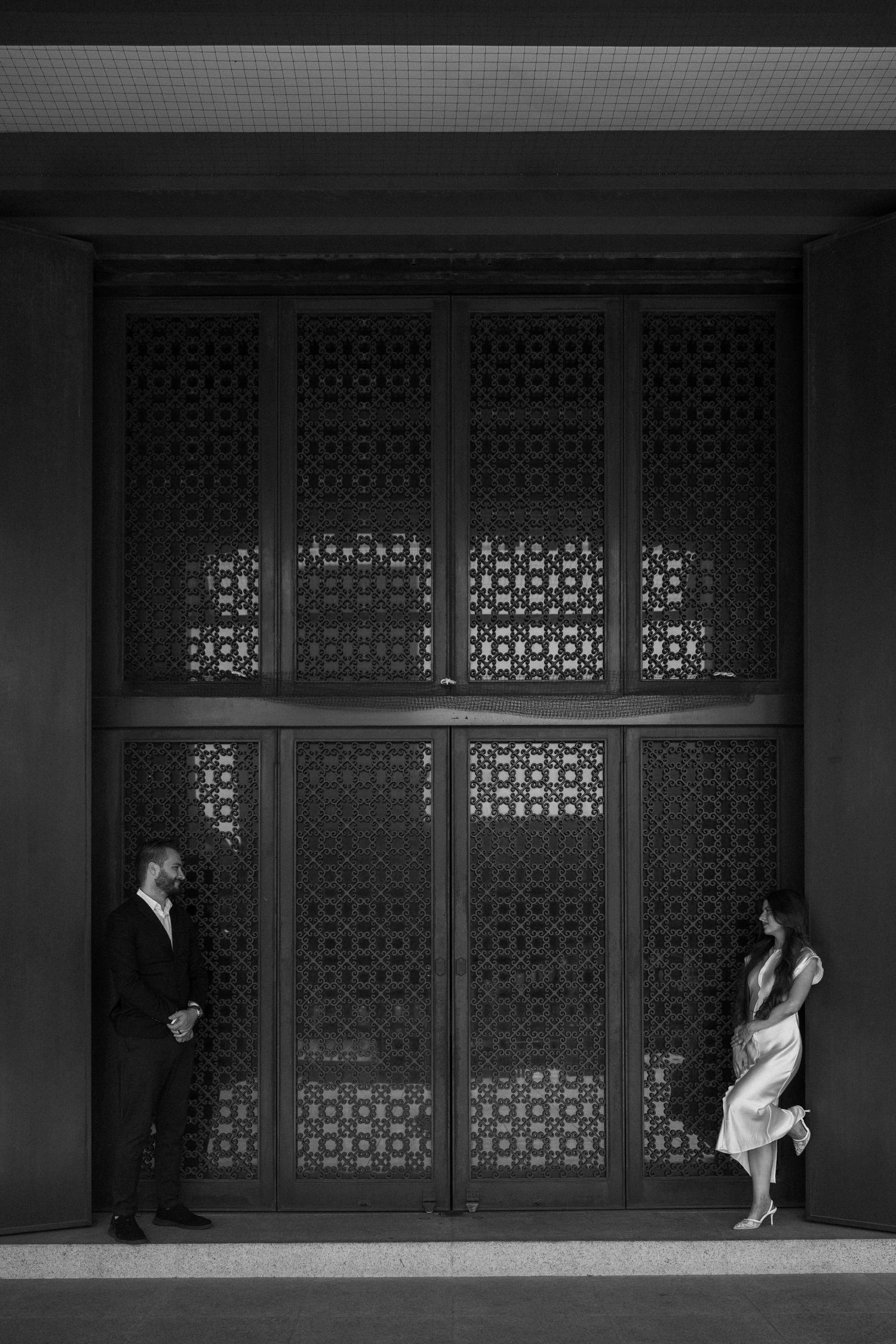Black and white photo of a couple standing apart by ornate latticed doors in elegant formal attire.