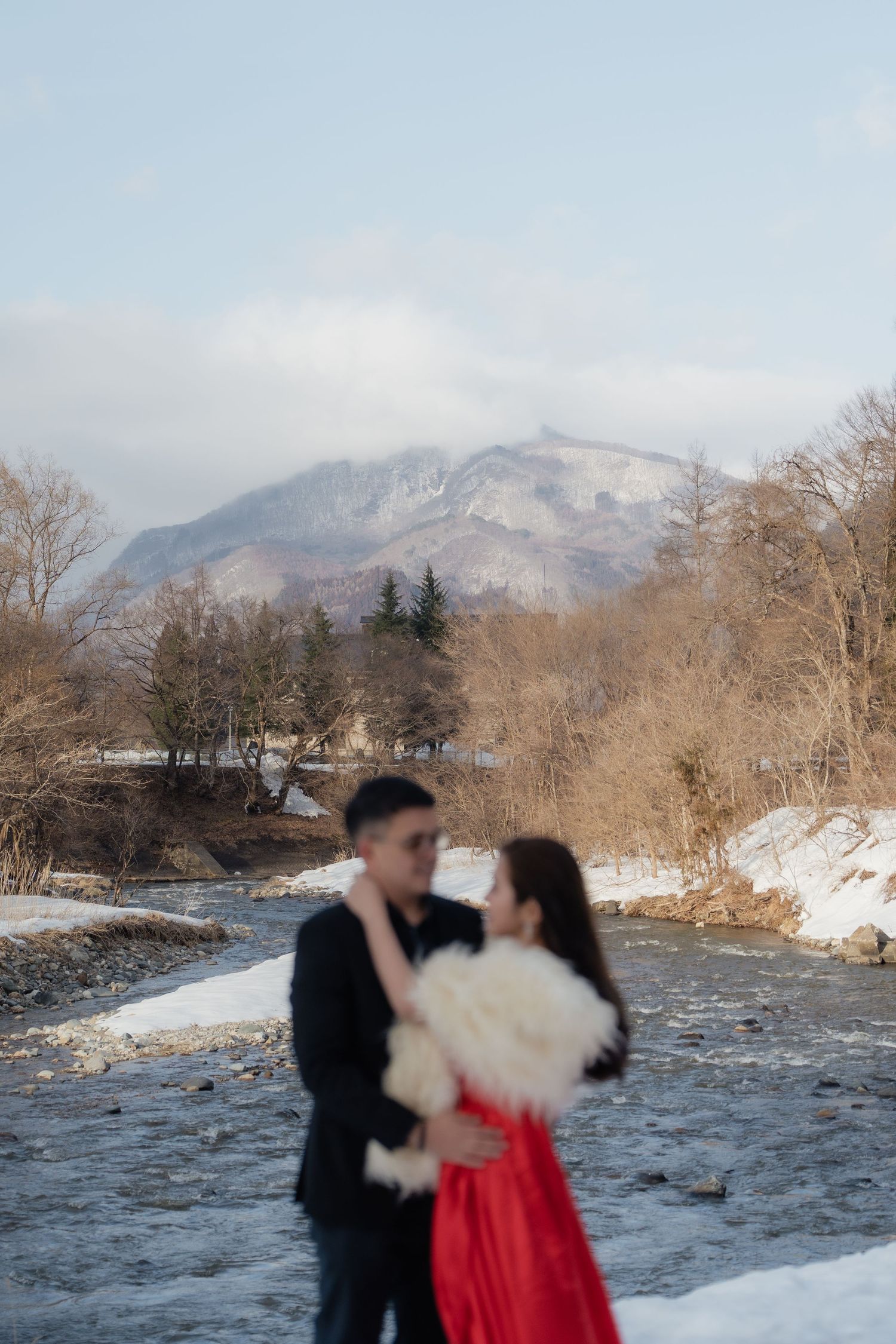 Couple embracing in winter landscape with snowy mountains and river, woman in red dress holding white bouquet.