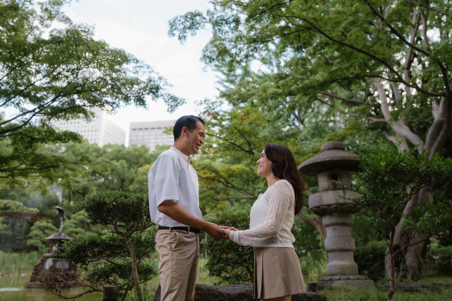 Couple holding hands and smiling at each other in a lush Japanese garden with stone lanterns.