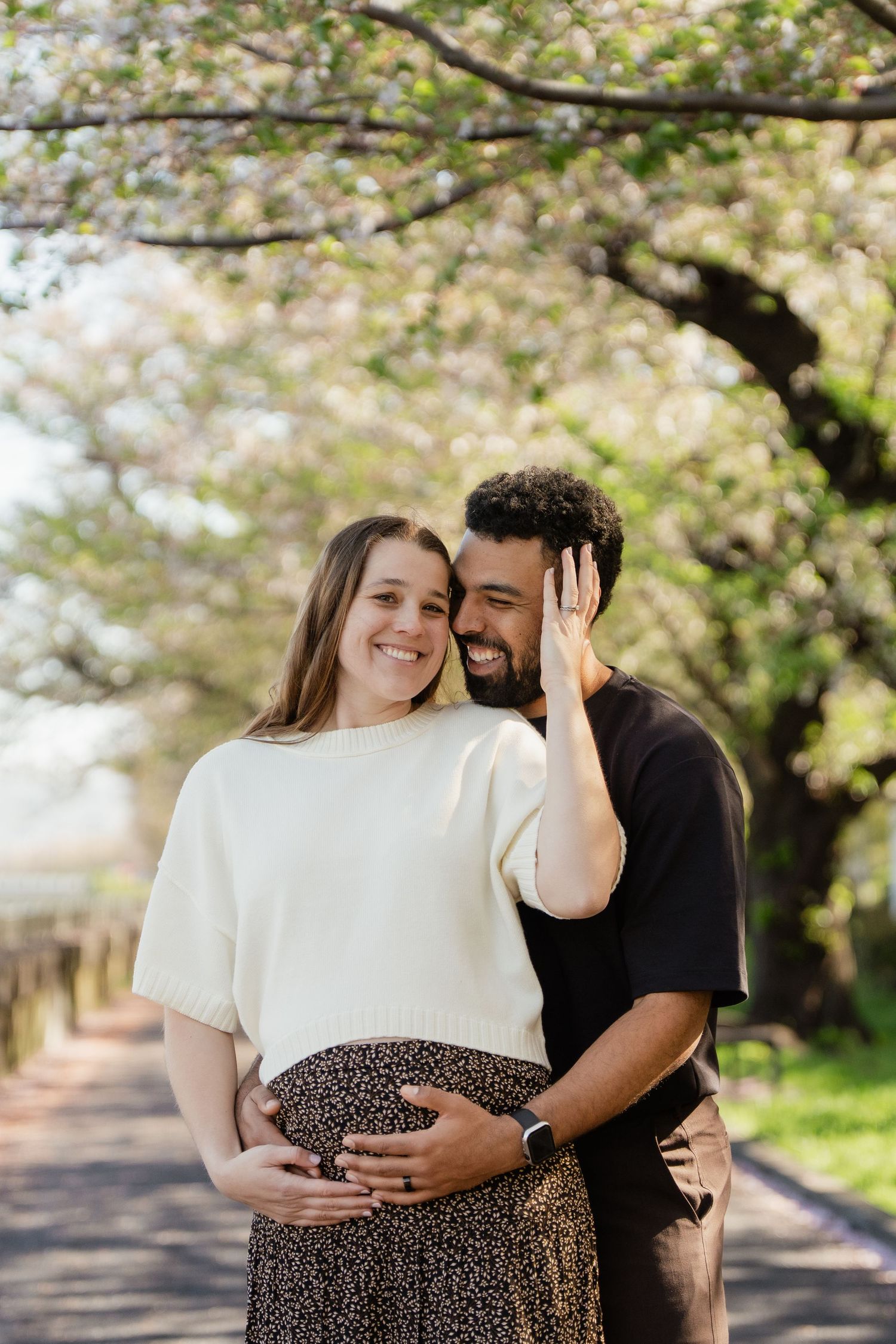 Happy couple embracing outdoors under blooming cherry blossom trees, woman in white top with baby bump, spring maternity photo.