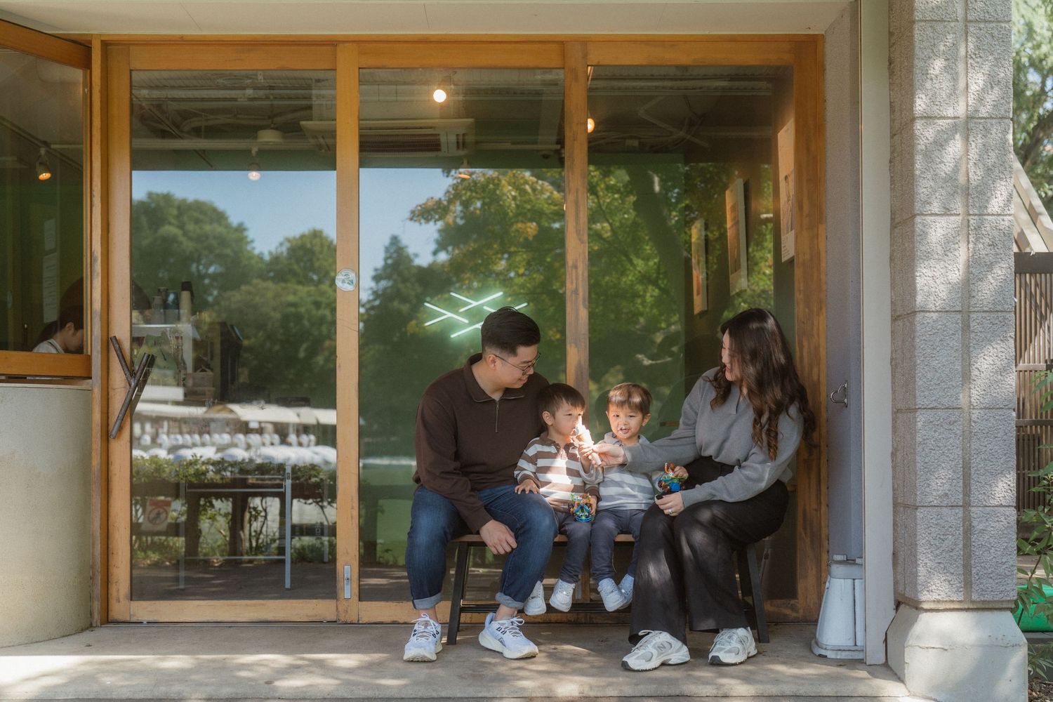 Family of four sitting together on a modern porch with large wooden-framed glass doors and lush green trees in background.