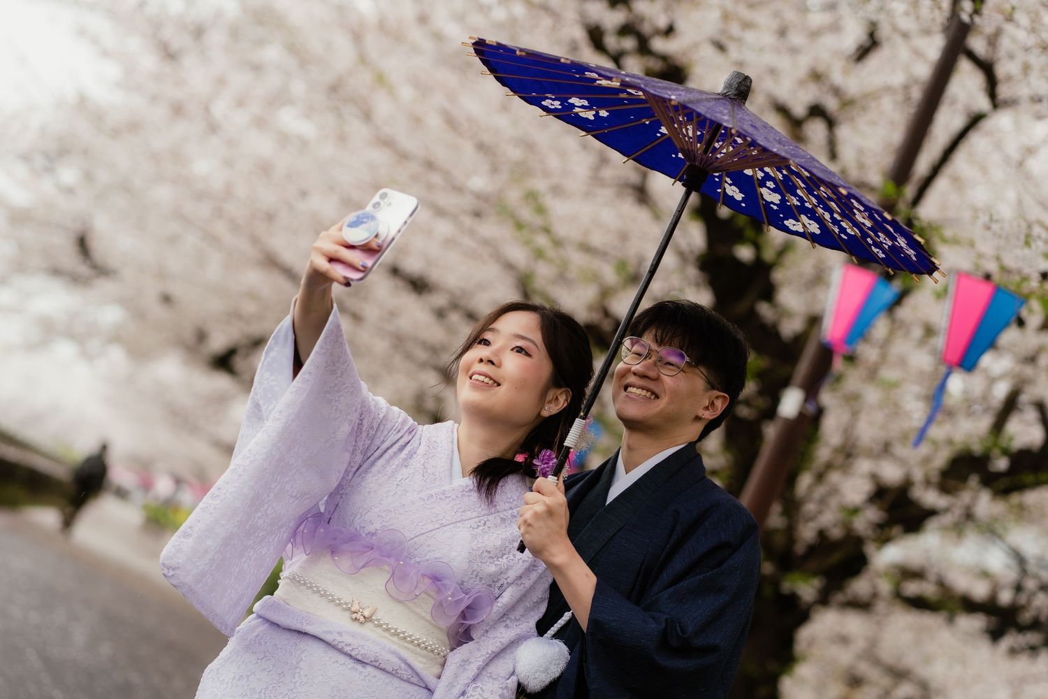 Couple in traditional Japanese kimono taking a selfie under a purple parasol with cherry blossoms in background.