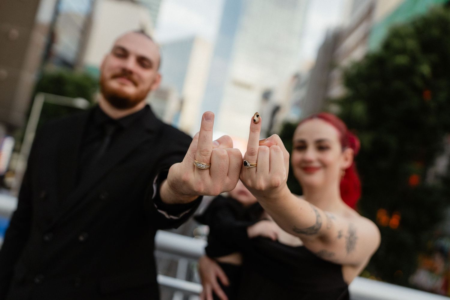 Couple in black outfits showing off wedding rings with middle fingers raised, city backdrop, tattooed woman with red hair.