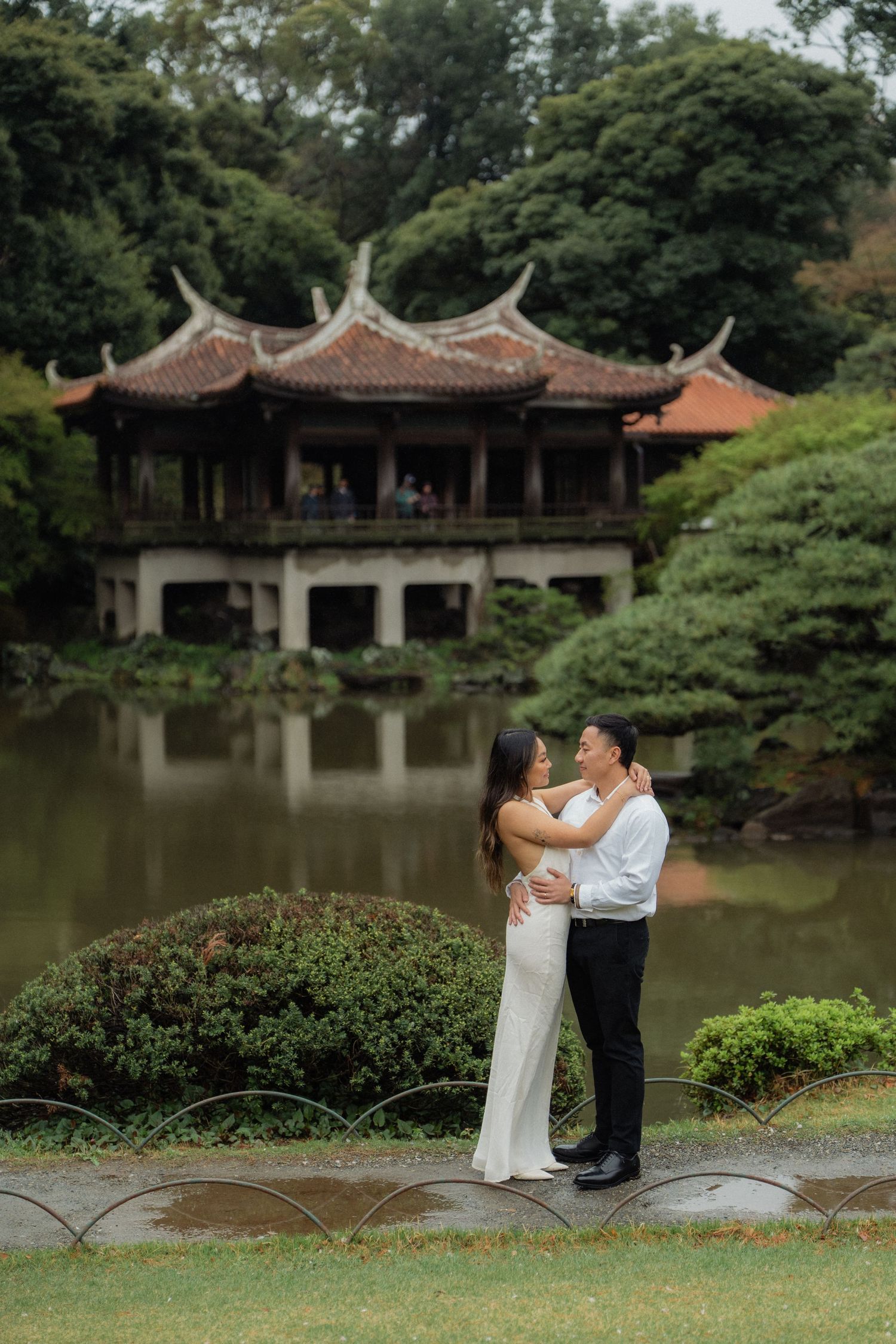 Couple embracing in white dress and black suit before a serene Japanese garden pond with traditional pagoda backdrop.
