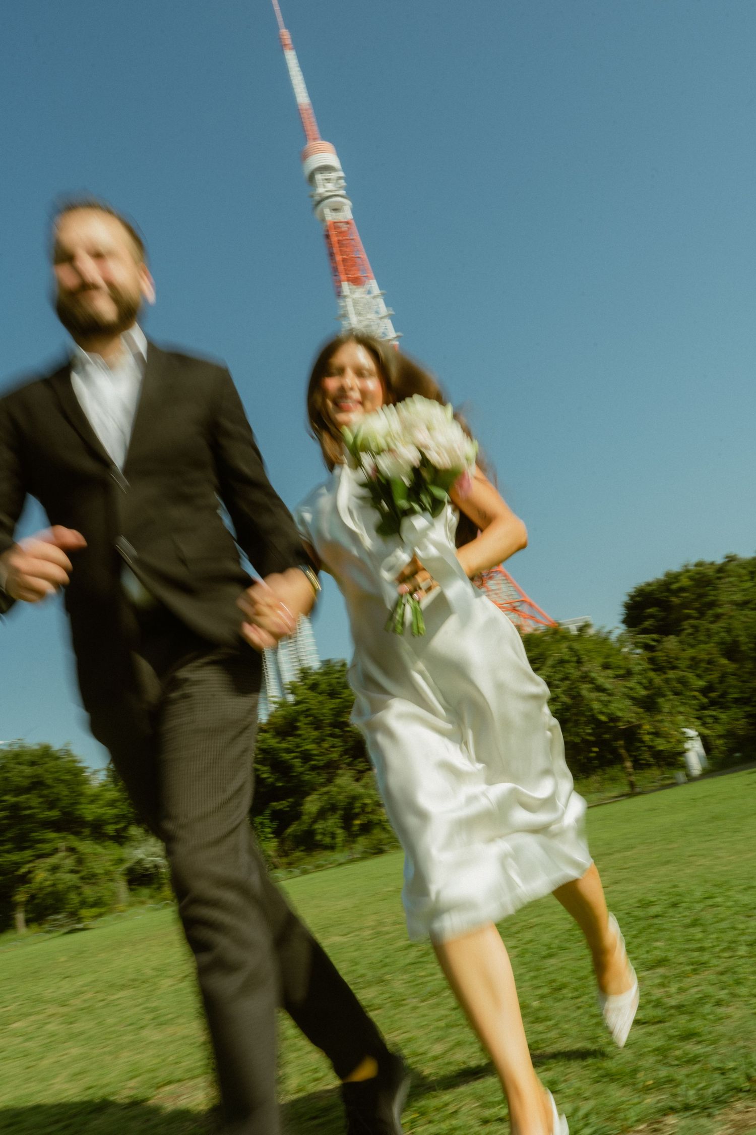 A joyful couple in wedding attire runs through a green park with Tokyo Tower visible in the background on a sunny day.