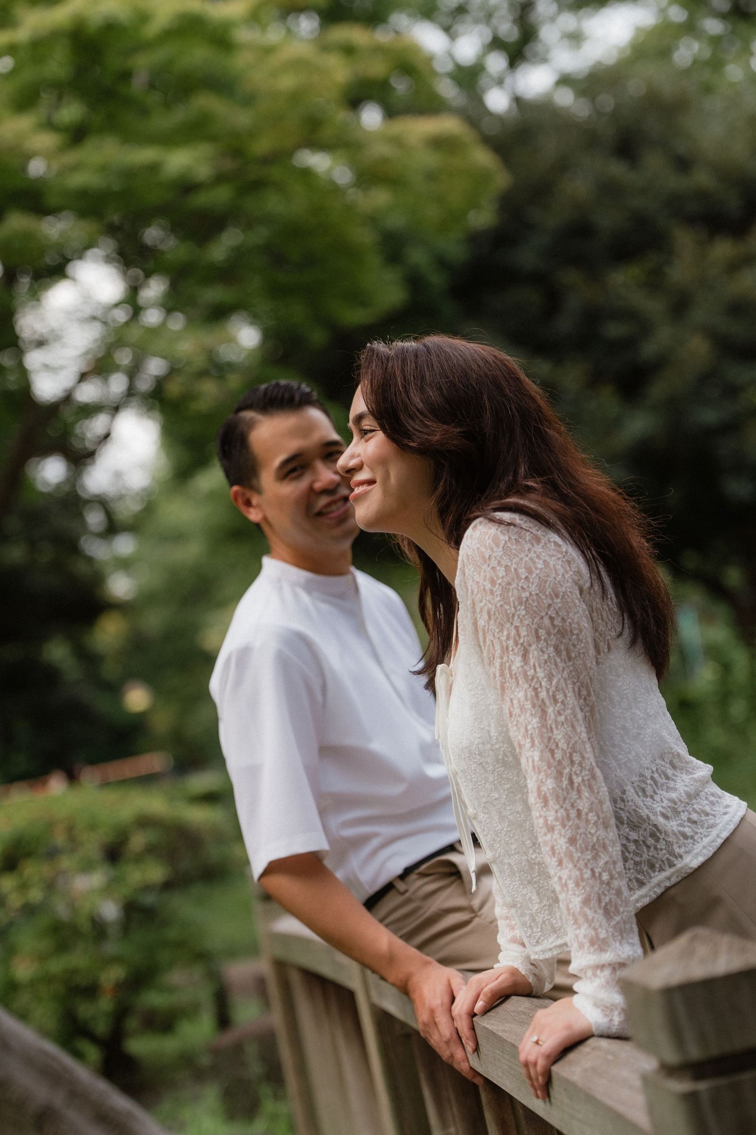 A happy couple shares a tender moment on a wooden bridge surrounded by lush green trees in a park.