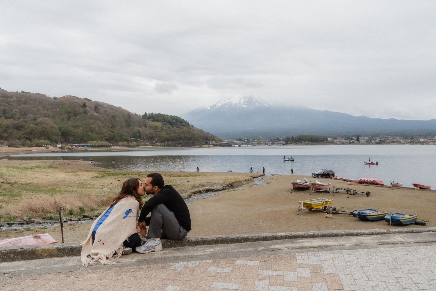 Couple sharing a romantic moment by Lake Kawaguchiko with Mount Fuji in the background on a cloudy day.