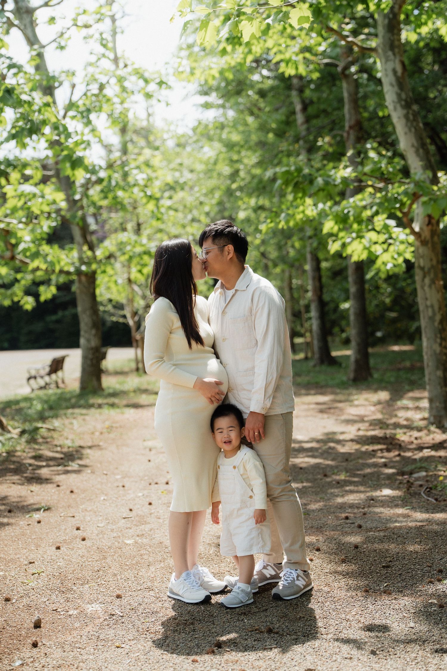 A happy Asian family of three poses on a park path, parents nearly kissing while toddler stands between them.
