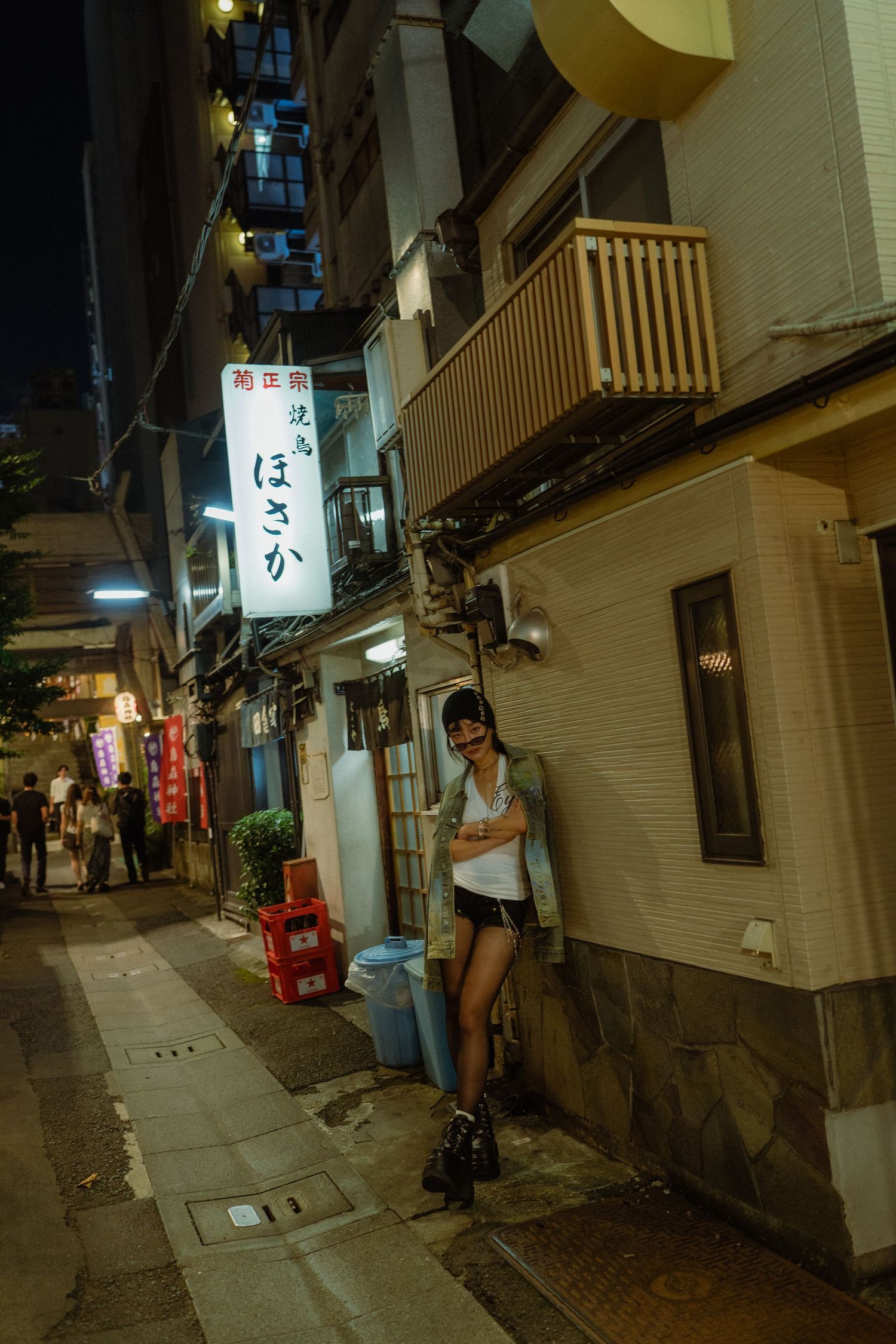 Young woman leaning against a building in a narrow Japanese alley at night, surrounded by illuminated kanji signs.