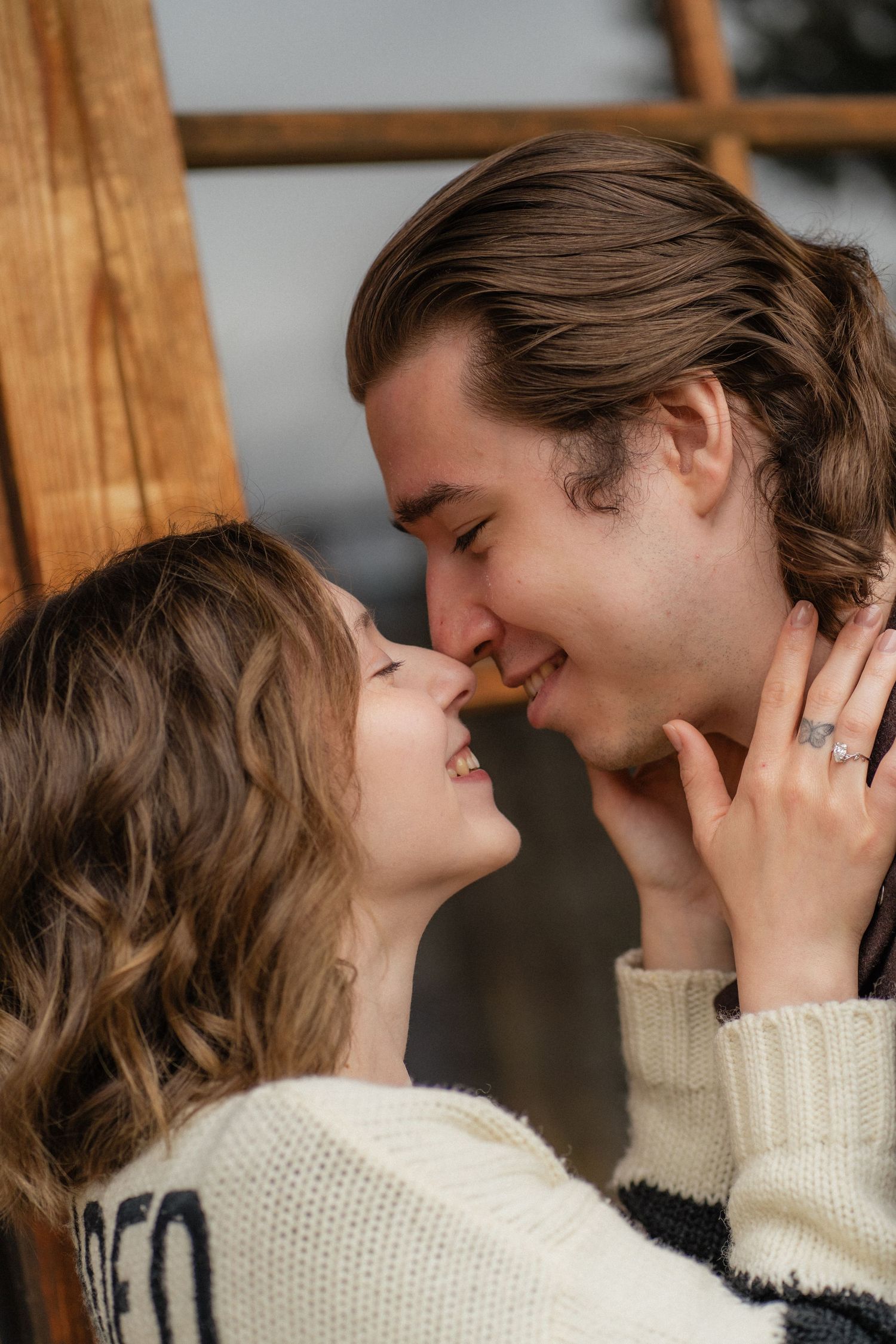 A smiling couple in cozy sweaters share a romantic moment, nearly kissing while holding each other tenderly indoors.