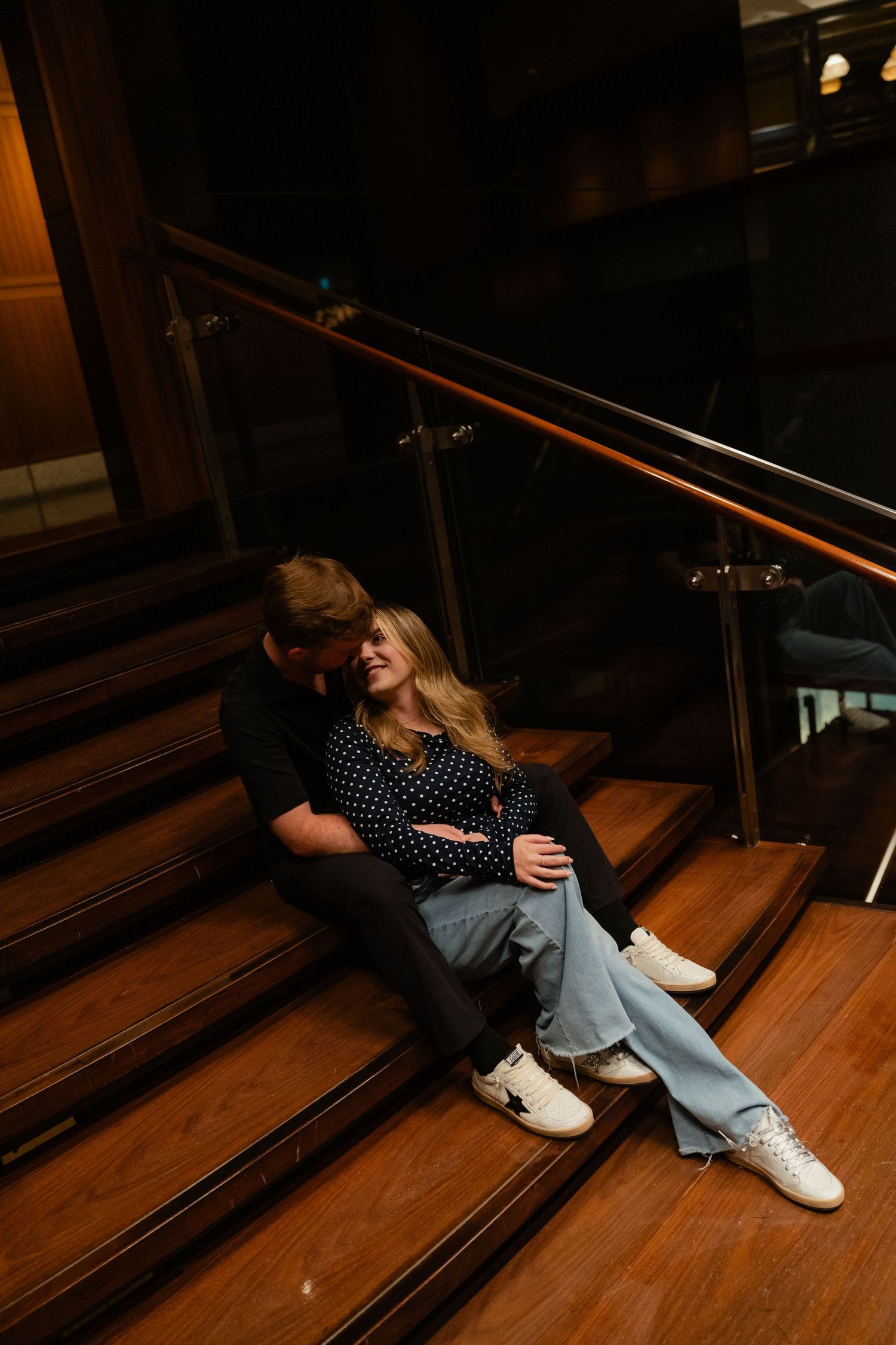 Couple sitting closely on wooden stairs with metal railing, woman in jeans and white sneakers leaning back smiling.