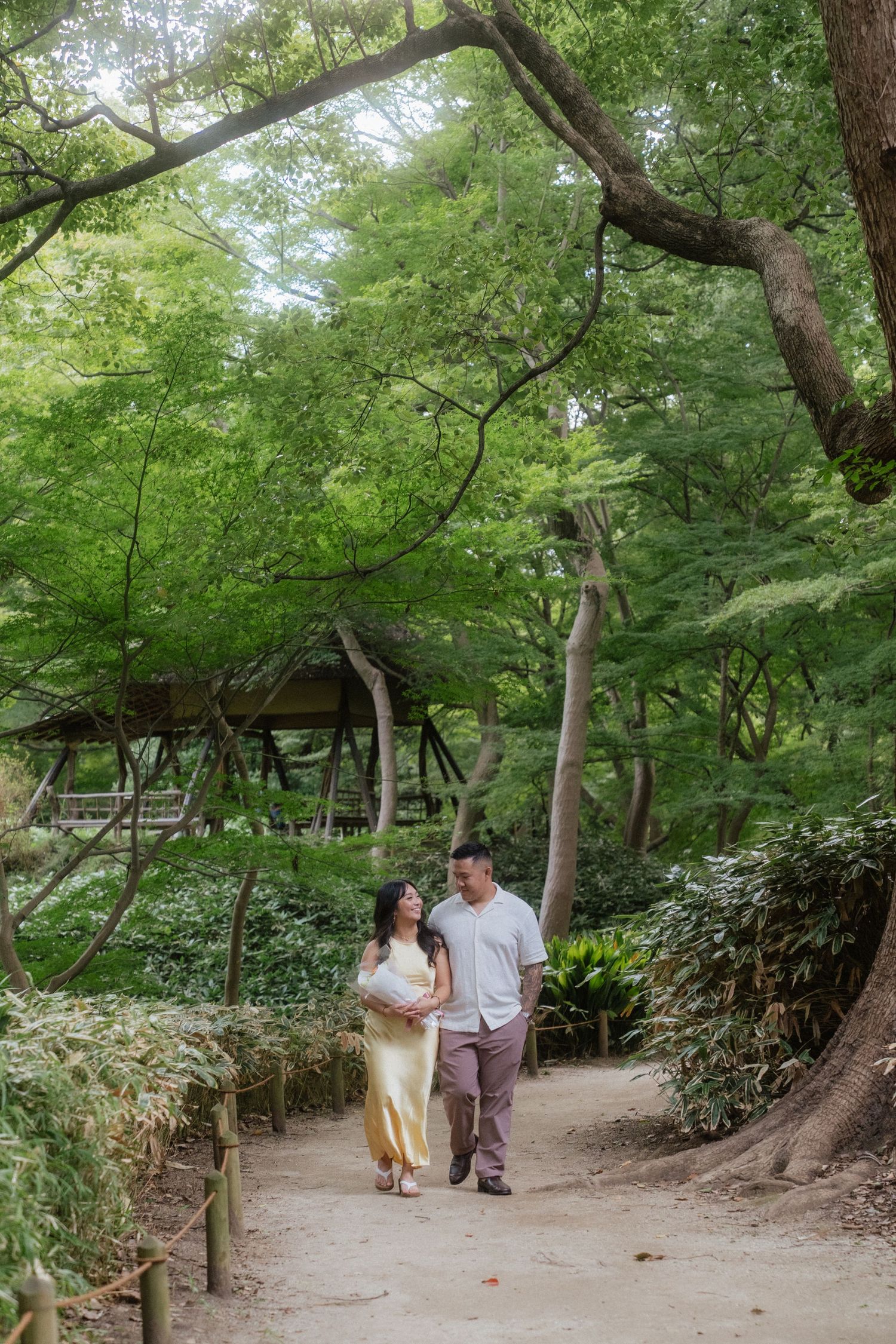 Couple walking hand in hand on a garden path surrounded by lush green trees in a serene park setting.