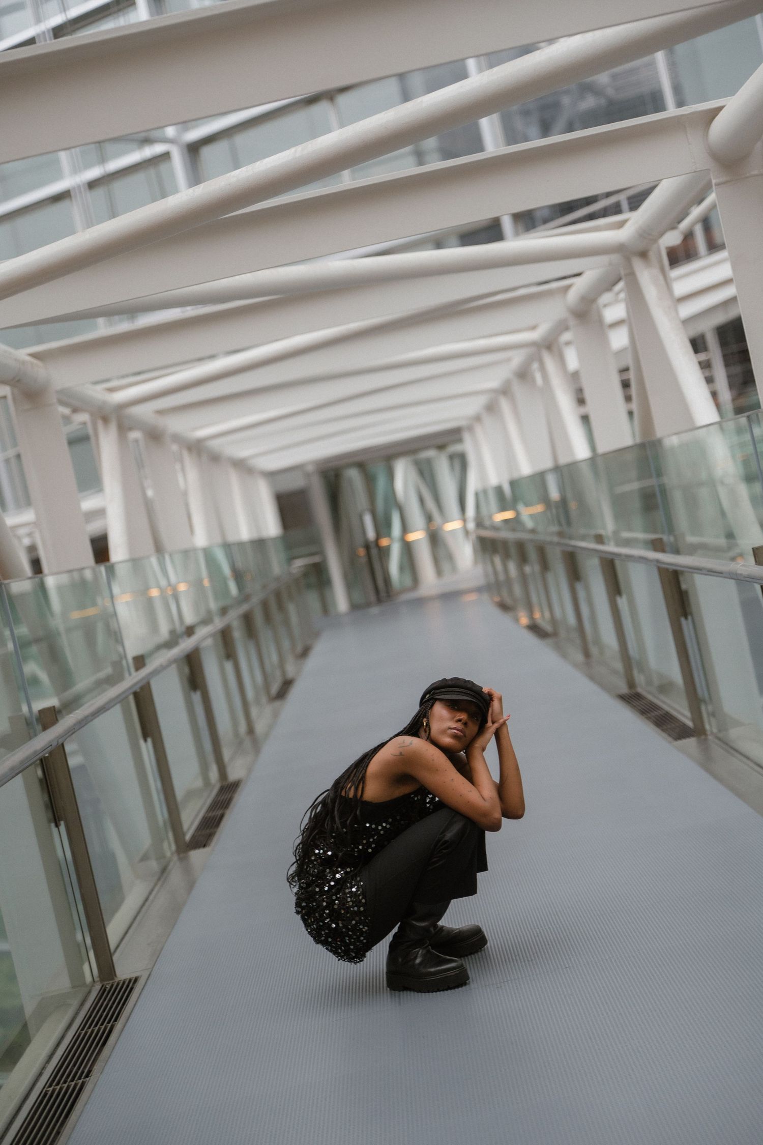 A young woman in black outfit crouches alone on a modern glass bridge corridor with white steel beams overhead.