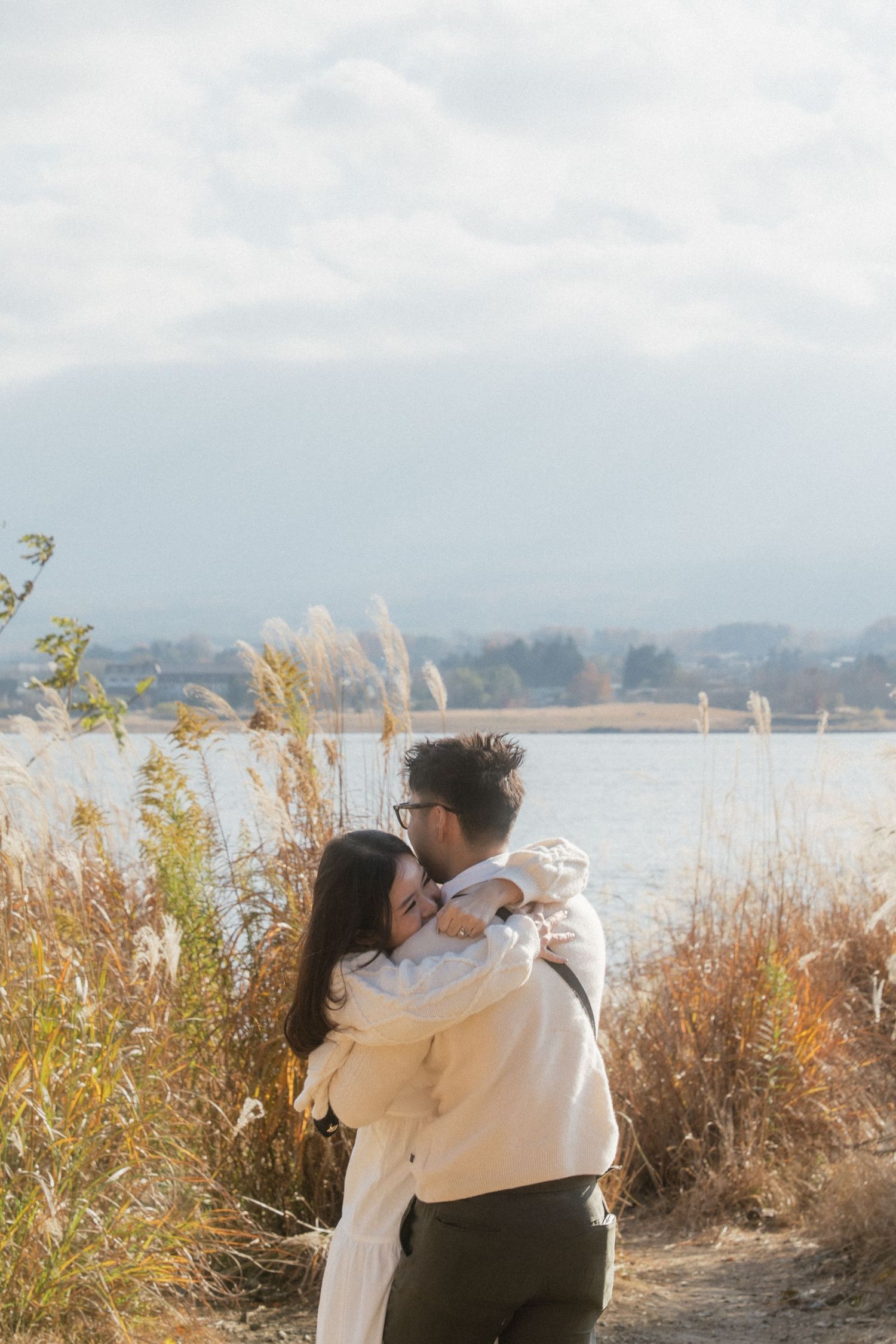 Couple embracing near a lake surrounded by golden tall grass on a cloudy autumn day.