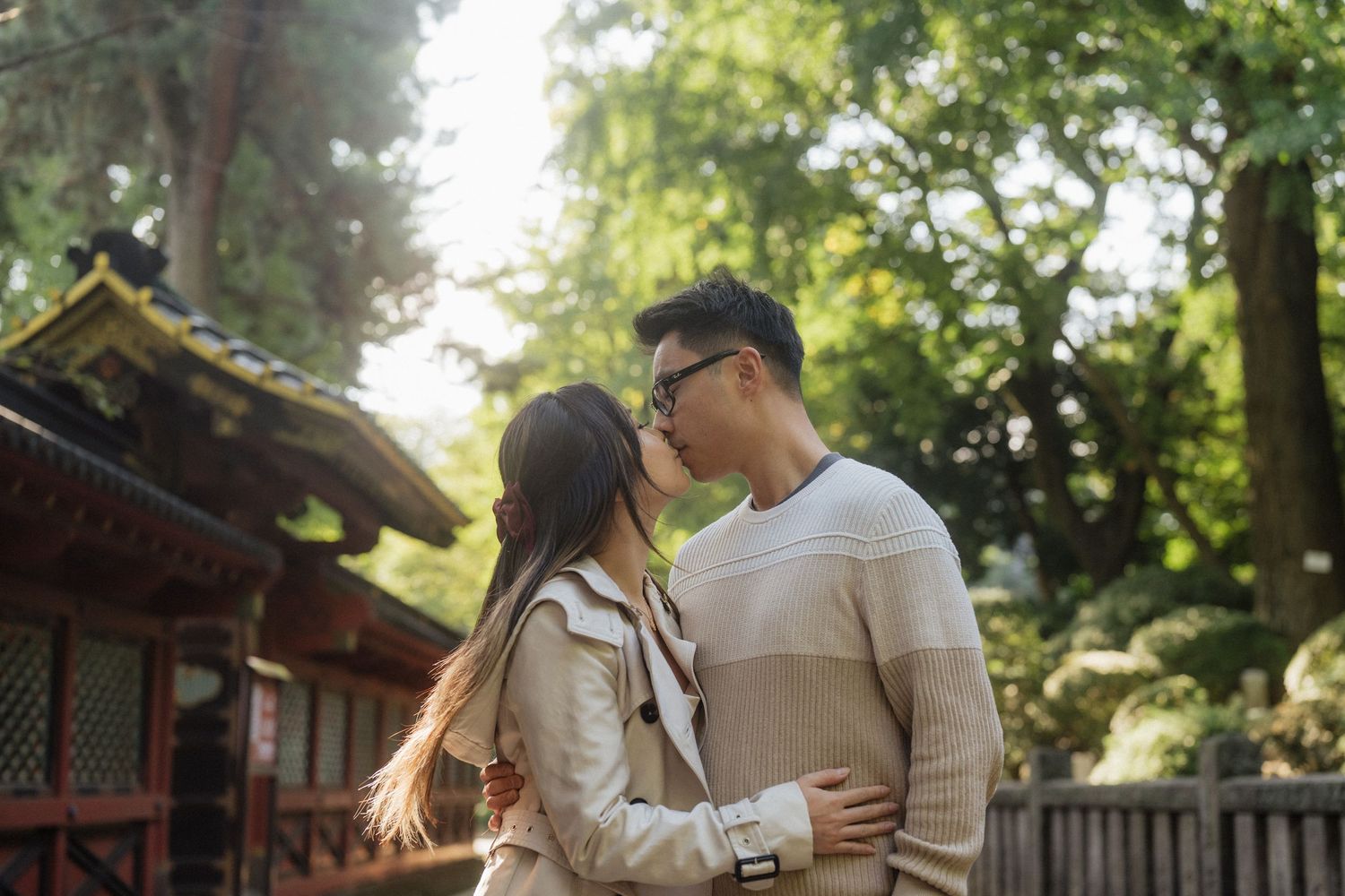 A couple shares a romantic kiss outdoors near a traditional Japanese building surrounded by lush green trees.