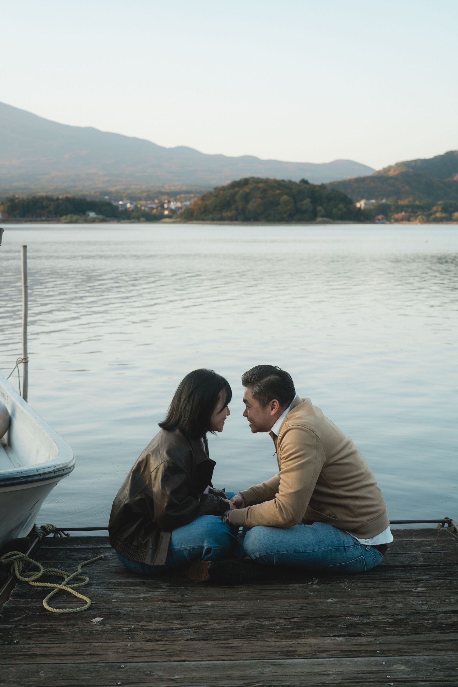 Couple sitting face-to-face on a wooden dock by a serene lake, holding hands with mountains in the background.