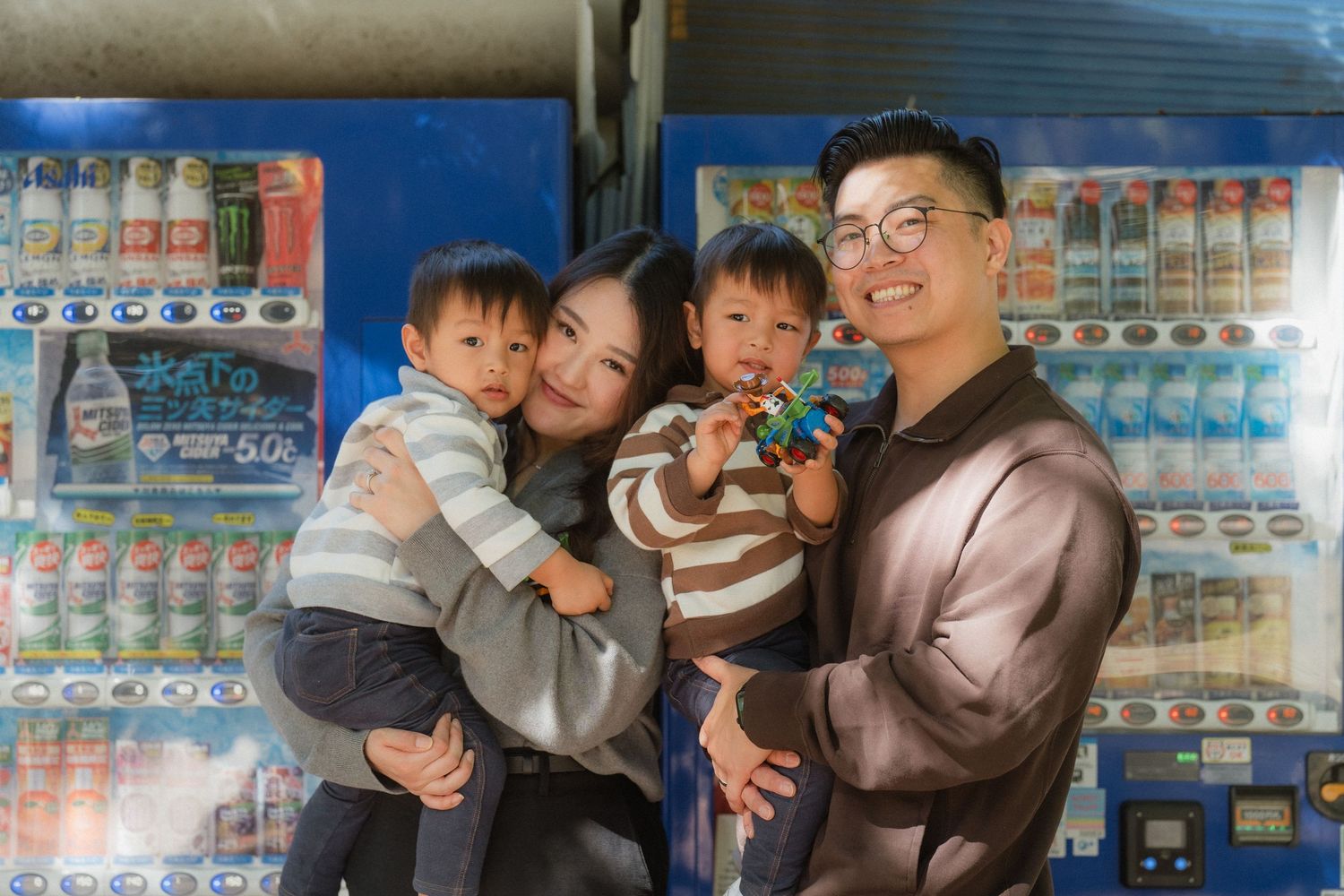 Happy Asian family with two toddlers posing in front of colorful vending machines outdoors.