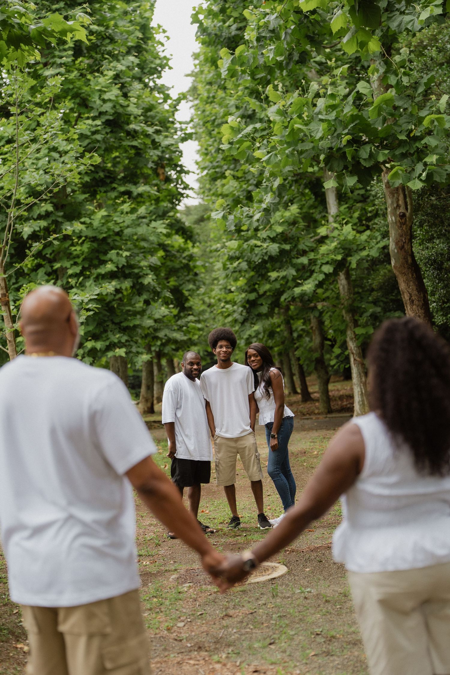 African American family holding hands in a tree-lined park path, with three smiling members posing in the background.