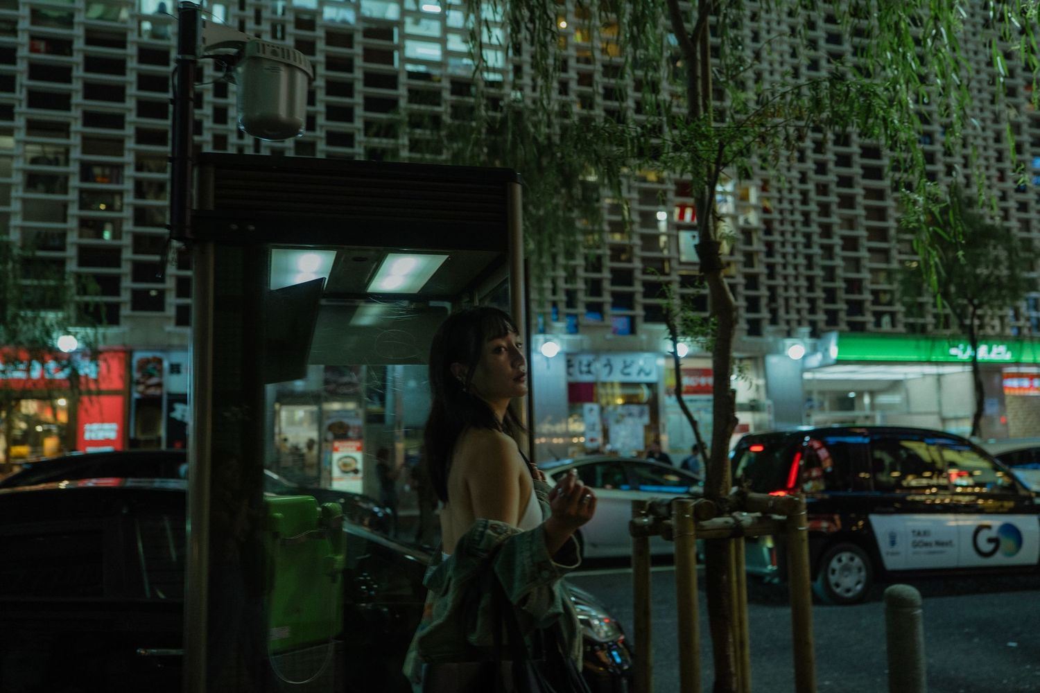 Woman standing at a city bus stop at night with urban buildings and green neon lights in the background.