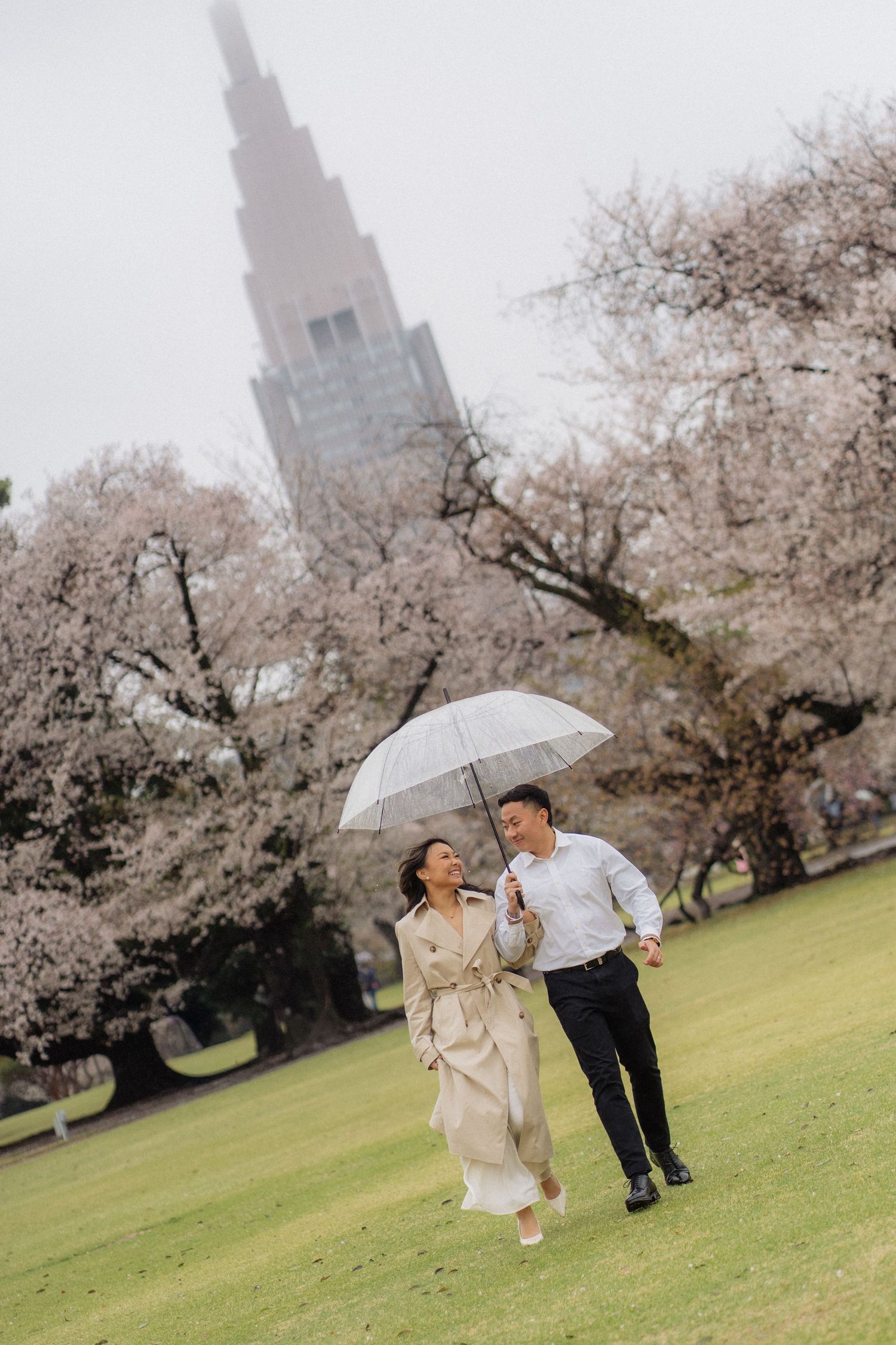 Couple walking under umbrella among cherry blossoms in Tokyo park with skyscraper backdrop.