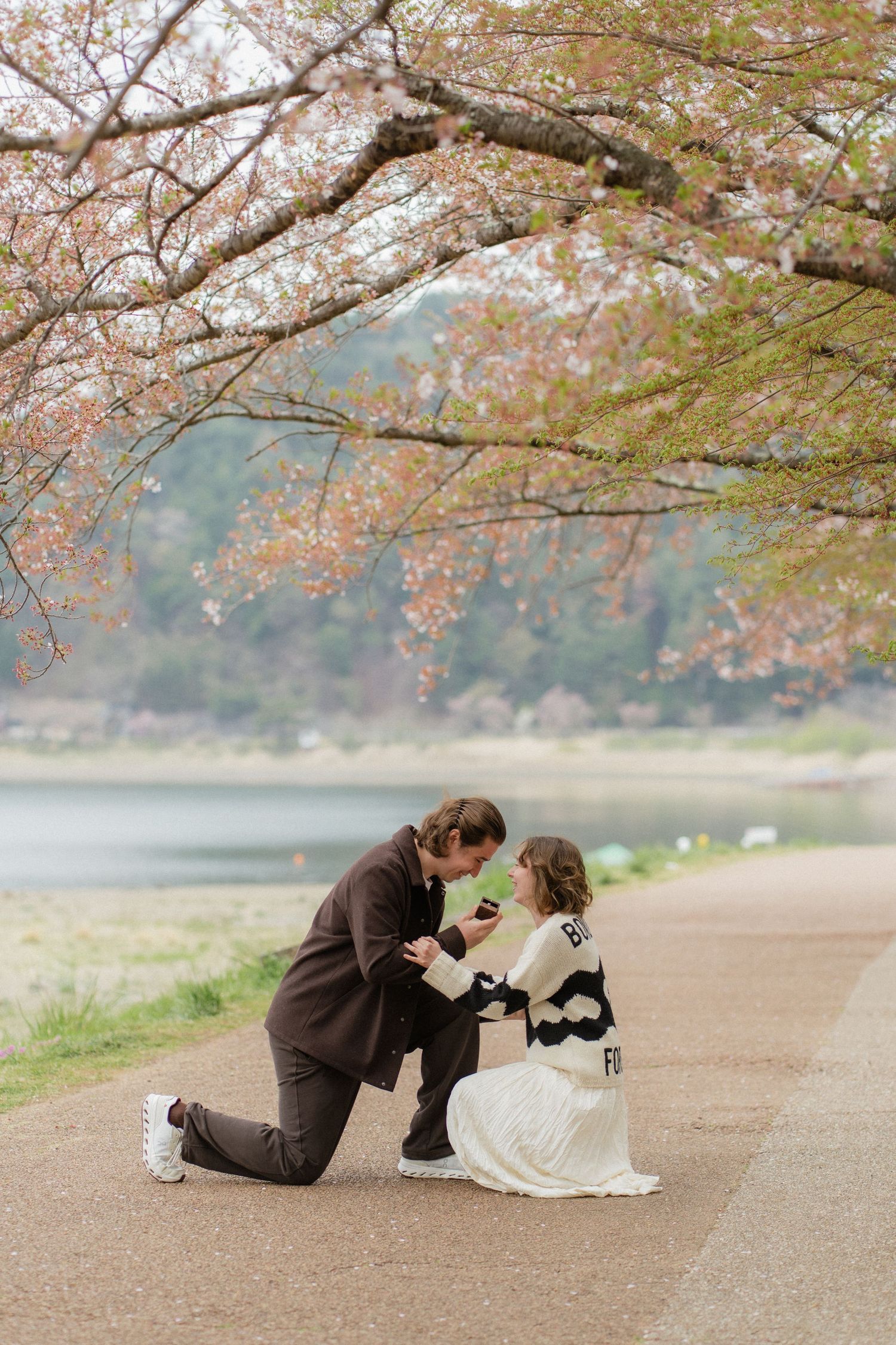 Couple sharing a romantic proposal moment under blooming cherry blossom trees beside a serene lake in spring.