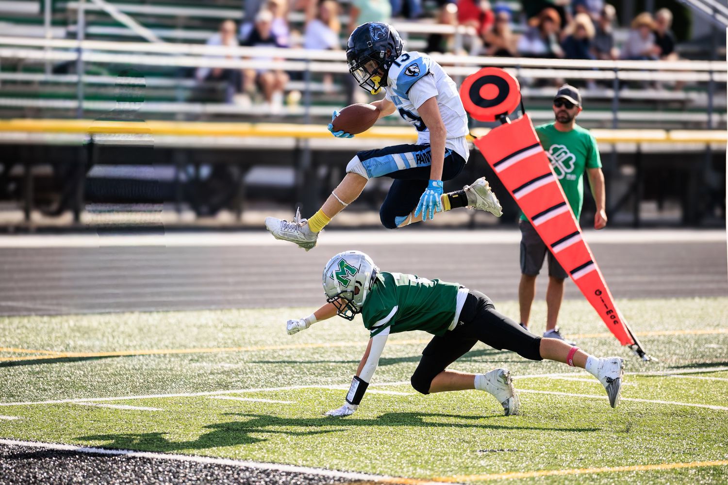 Football player in white leaps over diving defender in green during an outdoor game near the end zone marker.