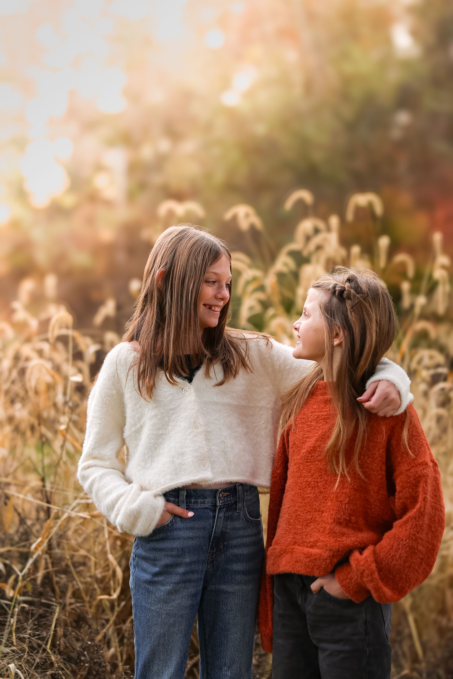 Two teenage girls smiling at each other in autumn field wearing cozy sweaters.