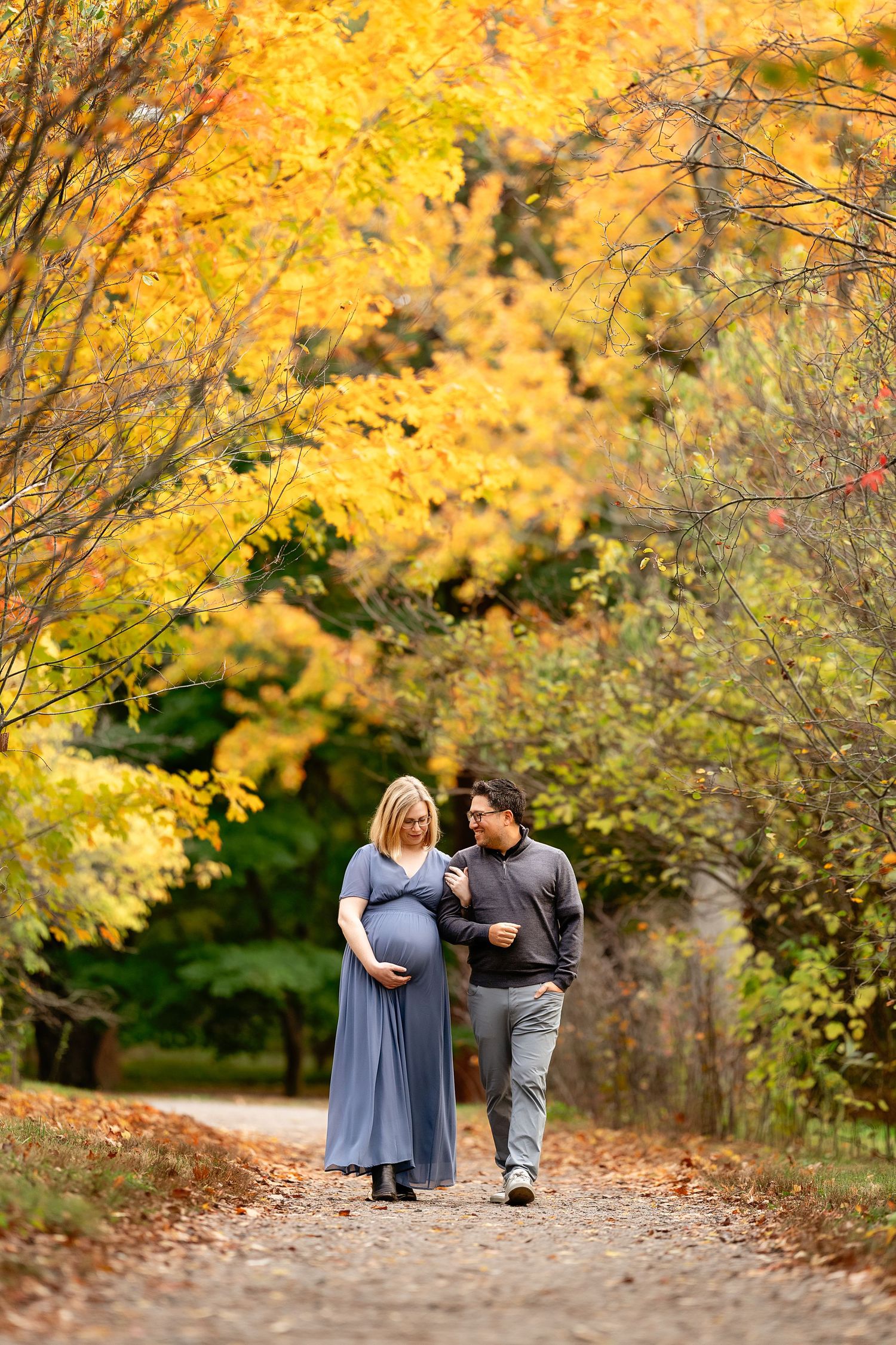 Expecting couple in a blue maternity gown and gray sweater walk arm in arm along a gravel path beneath golden autumn trees.