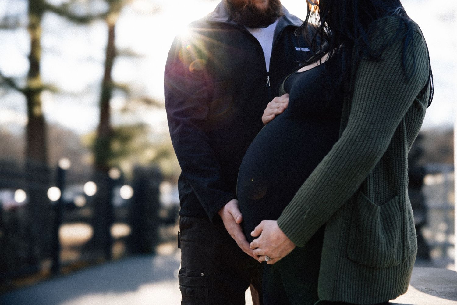 A couple cradles a pregnant belly together outdoors in soft sunlight during a maternity shoot.