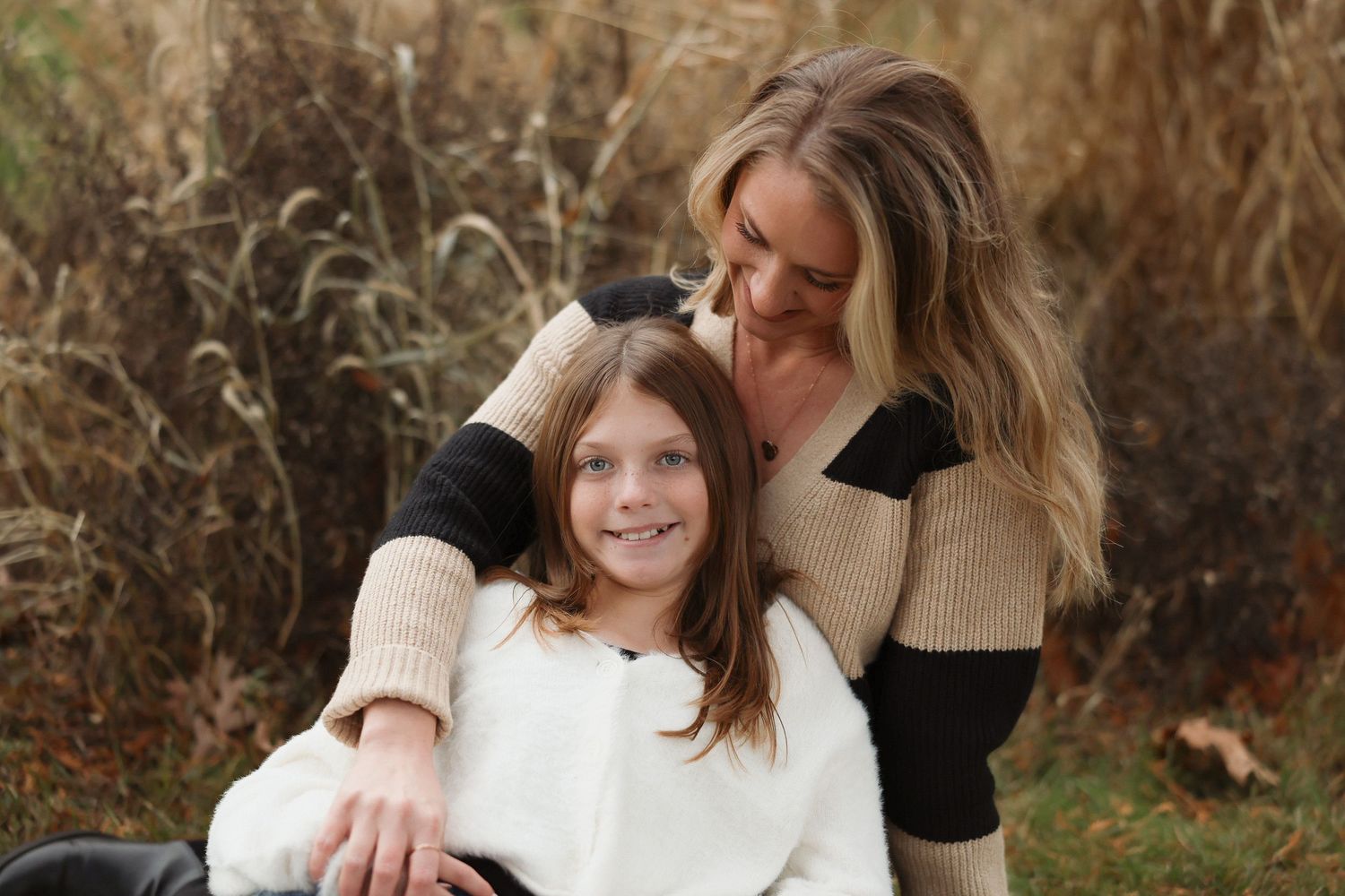 A mother and daughter smile together outdoors in a fall field, wearing cozy sweaters.
