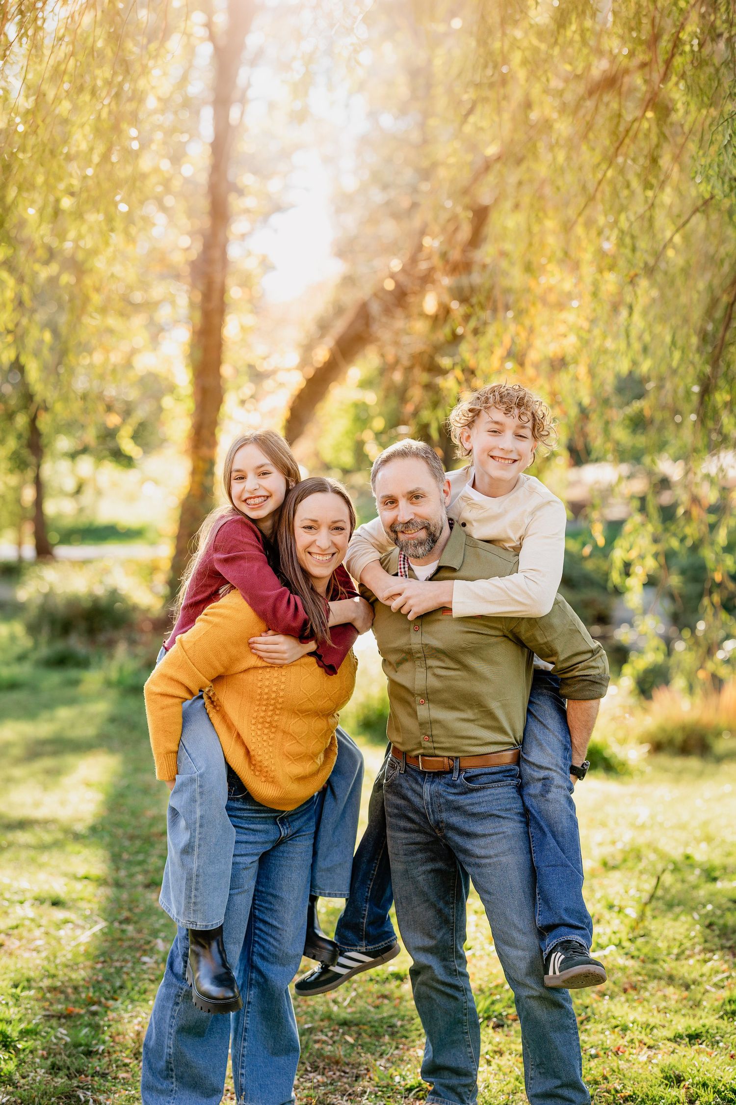 A smiling family of four poses outdoors in golden sunlight as the parents give their daughter and son piggyback rides under willow trees.