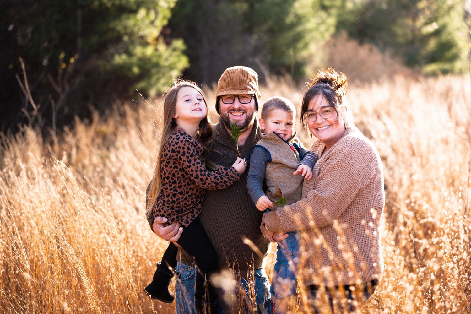 Happy family of four posing in golden autumn field, parents holding young children, smiling outdoors.