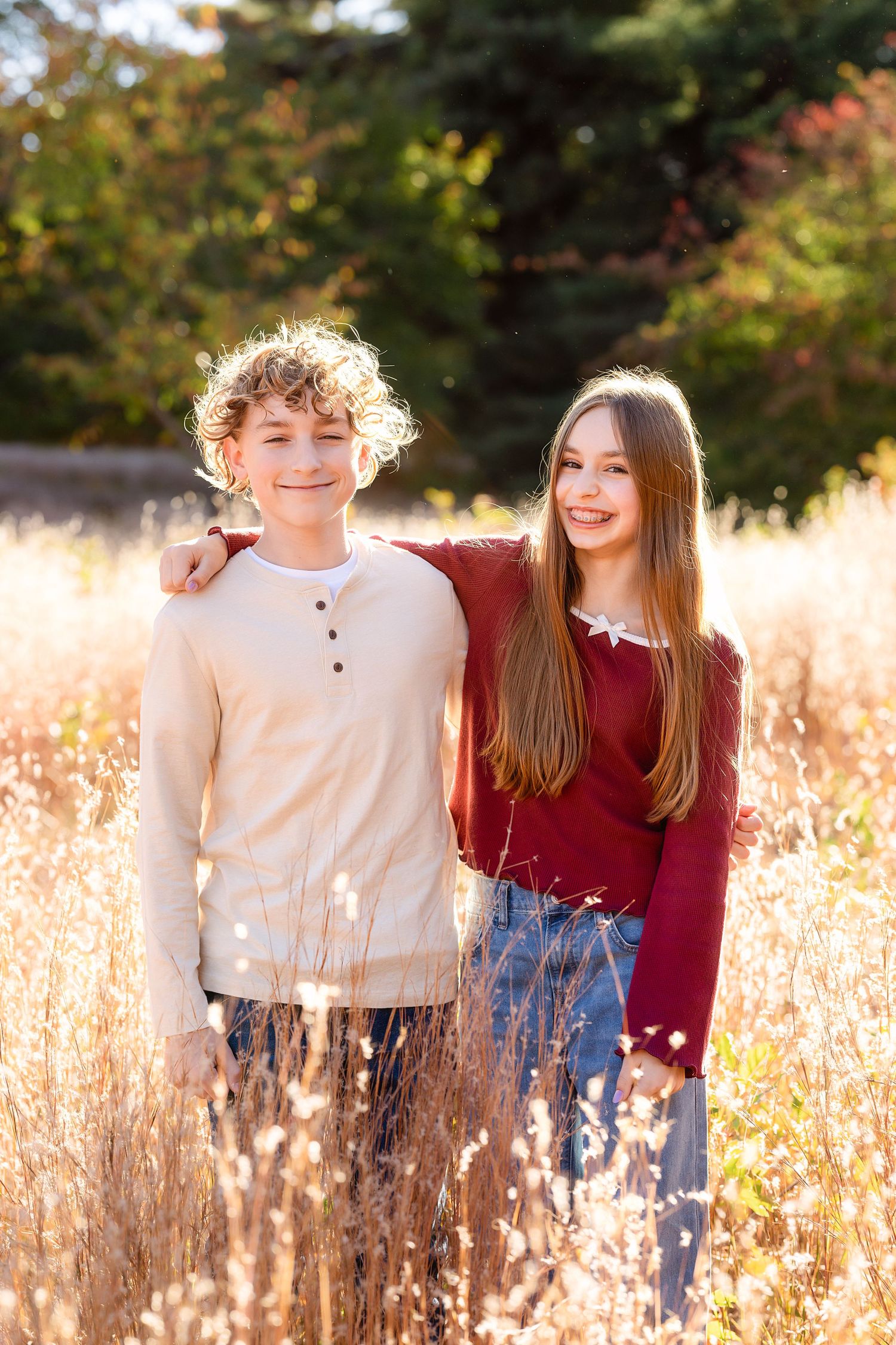 Two smiling siblings pose with arms around each other in a golden autumn field, wearing a cream henley and a maroon top.