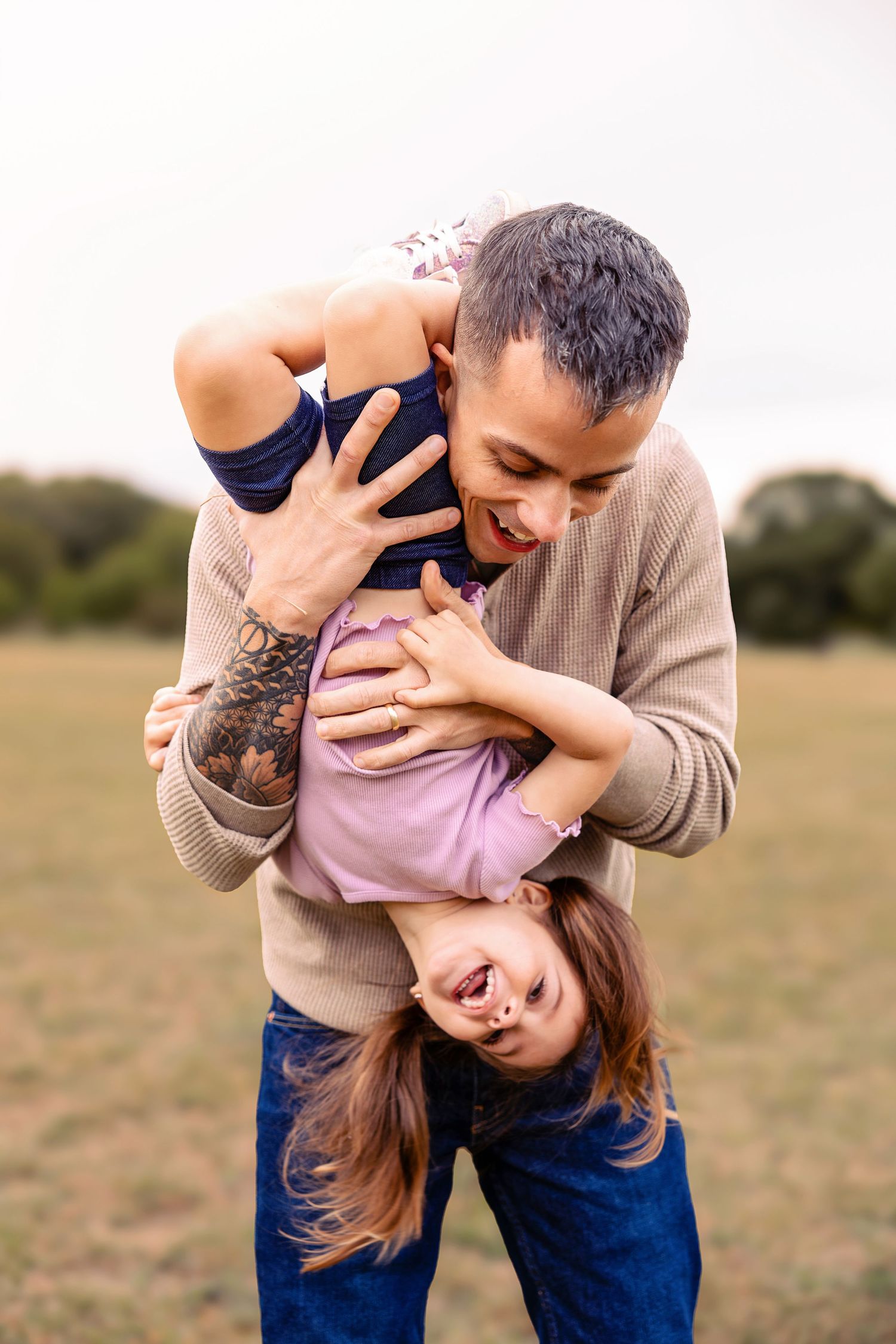 Smiling man with a tattooed arm playfully holds a laughing little girl upside down in an open grassy field on a bright, cloudy day.