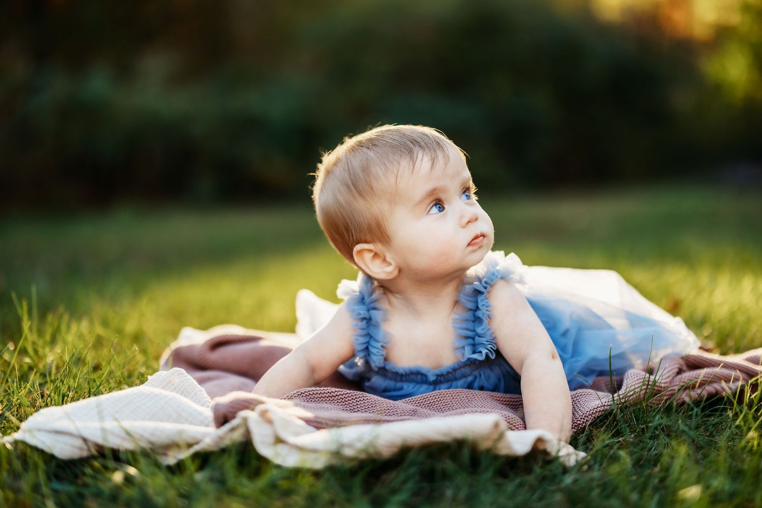 Baby in a blue ruffled outfit lies on a soft blanket in a sunlit green grassy park, looking curiously to the side.