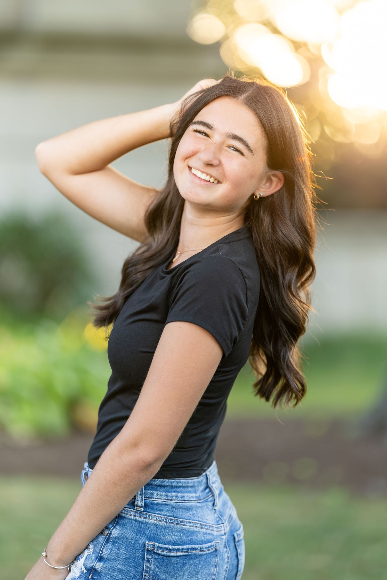 Smiling teenage girl with long dark hair in black shirt and denim shorts posing in golden sunlight.