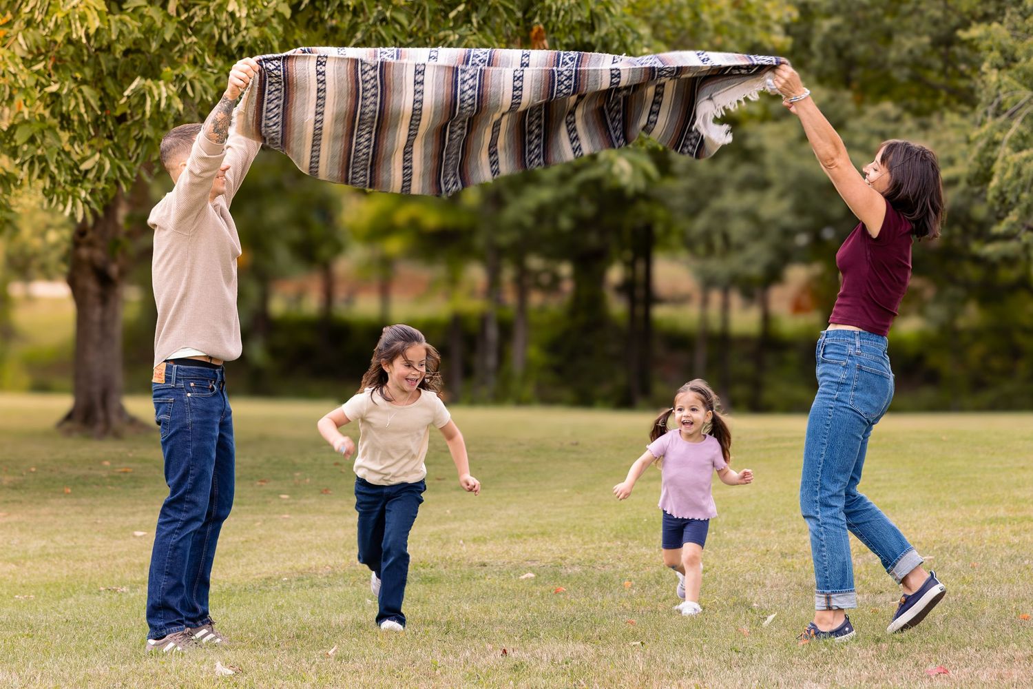 Family playing outdoors in park, parents holding blanket overhead while two young girls run underneath on green grass.