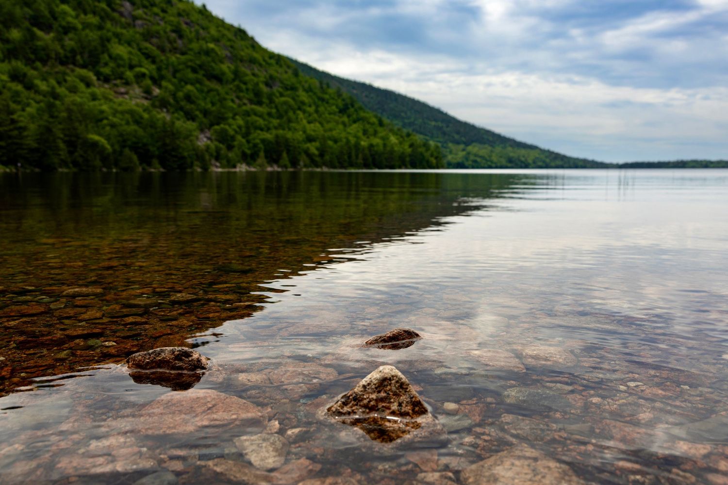 Tranquil lake with rocky shoreline reflecting green forested hills under a cloudy sky.