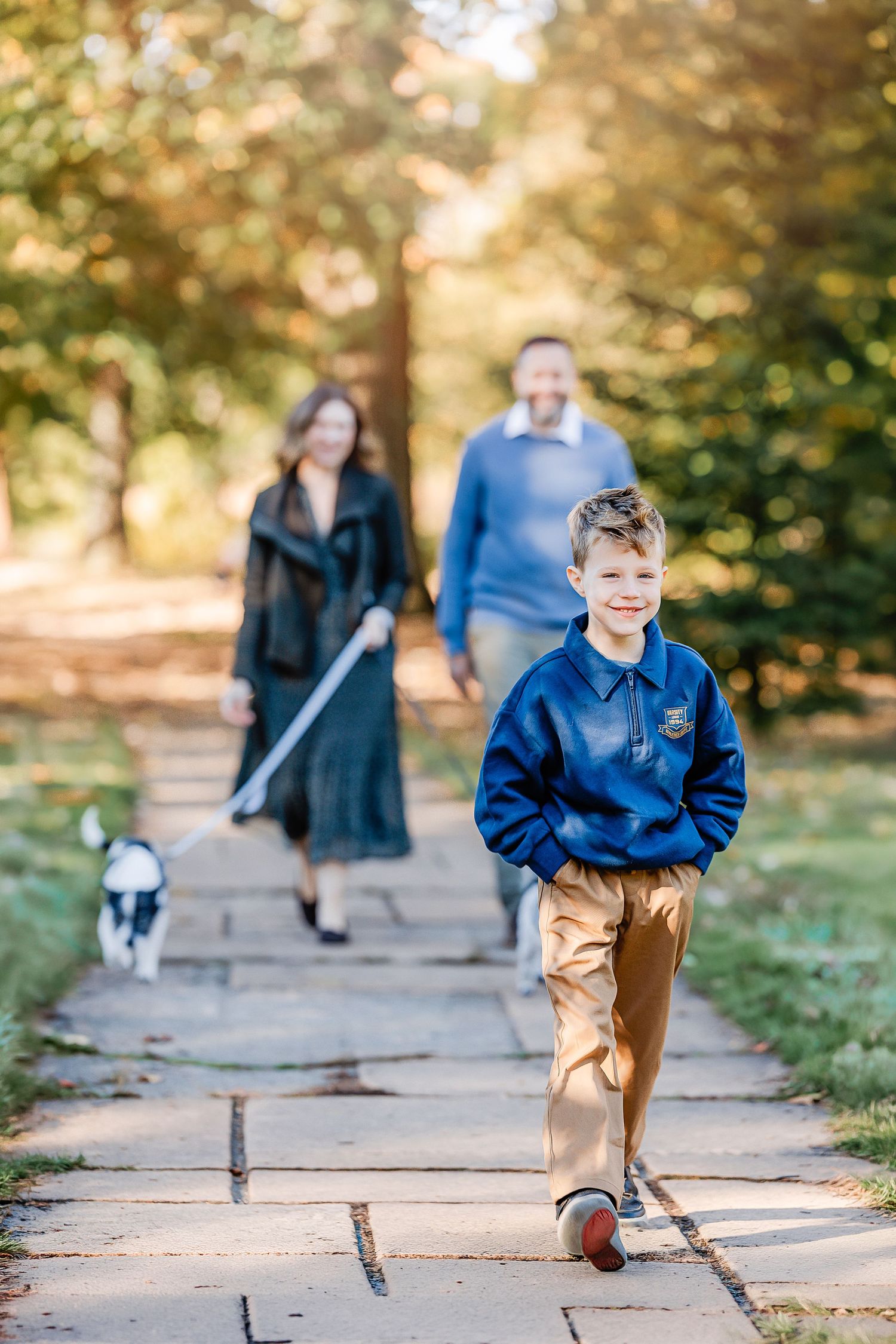 Smiling boy in a blue jacket and khaki pants walks along a stone path in a sunny park while his parents follow with a small dog.