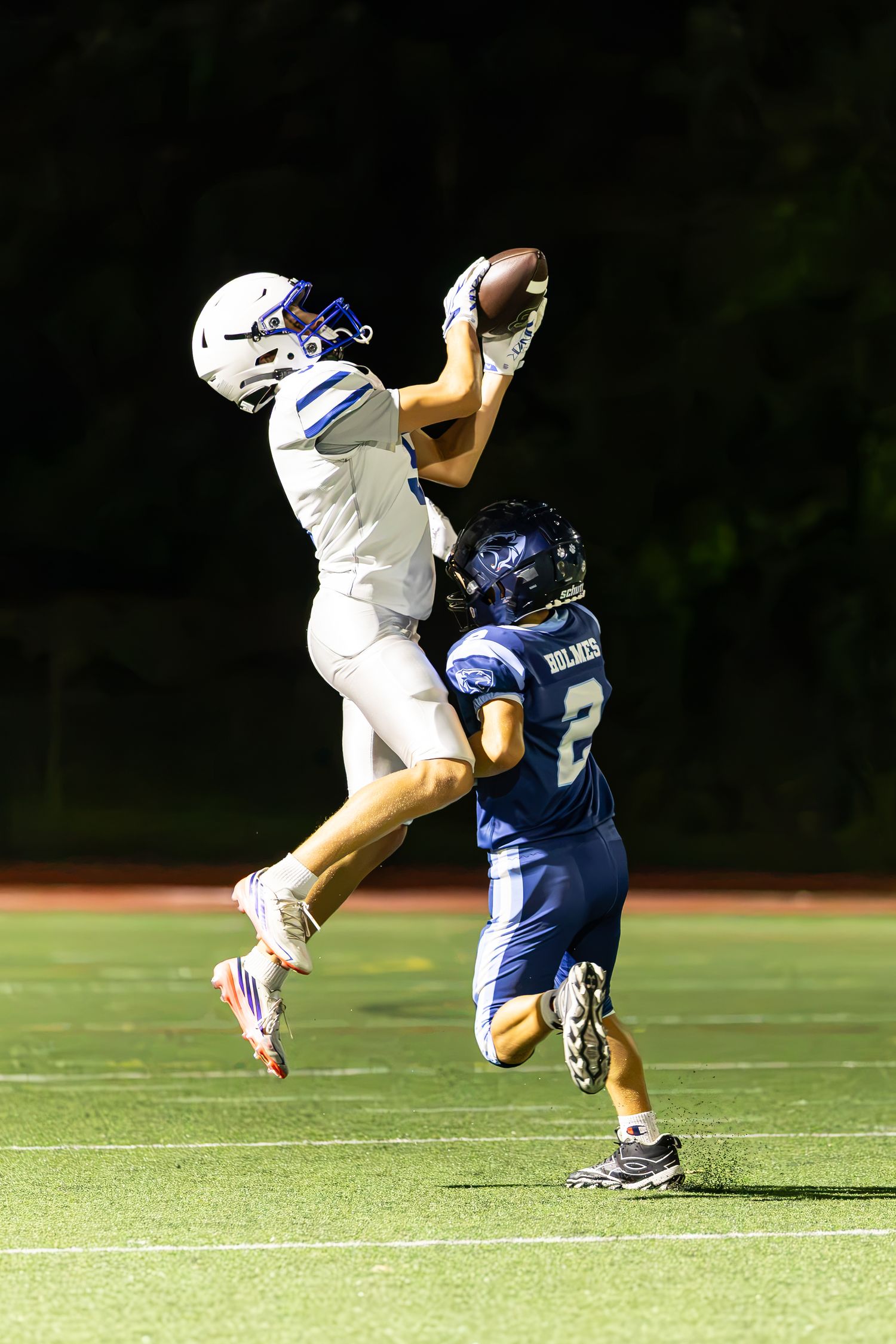 Football player in white uniform leaps to catch the ball over a defender wearing number 2 during a night game.