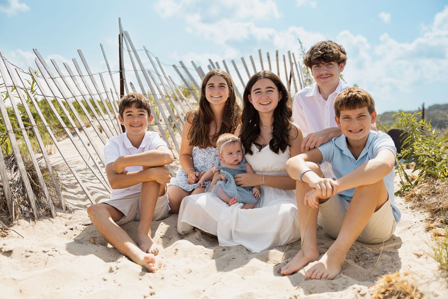 Group of five siblings sitting together on sandy beach with wooden fence backdrop on sunny day.