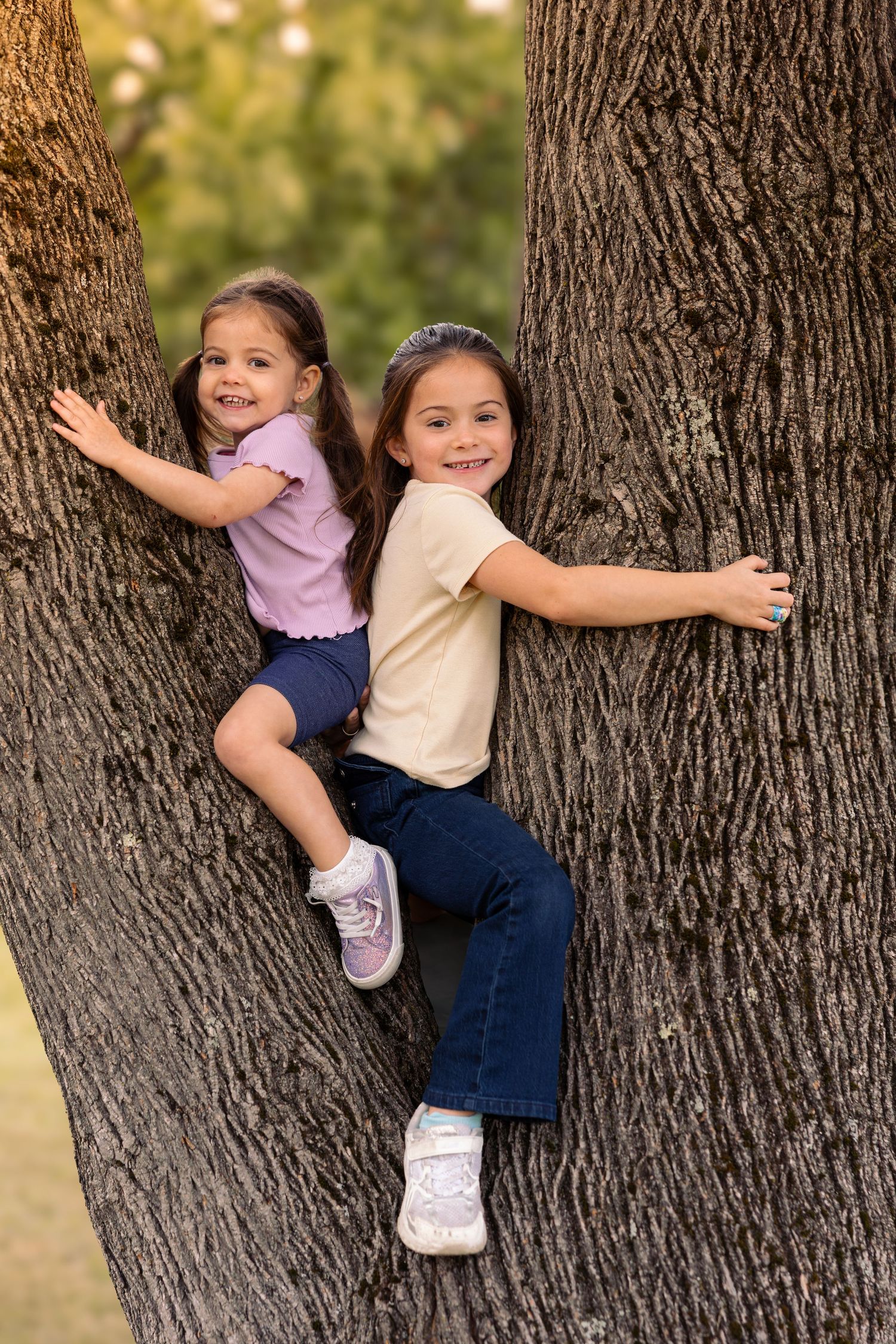 Two young girls smiling and hugging a large tree trunk in a park, wearing casual clothes and sneakers.