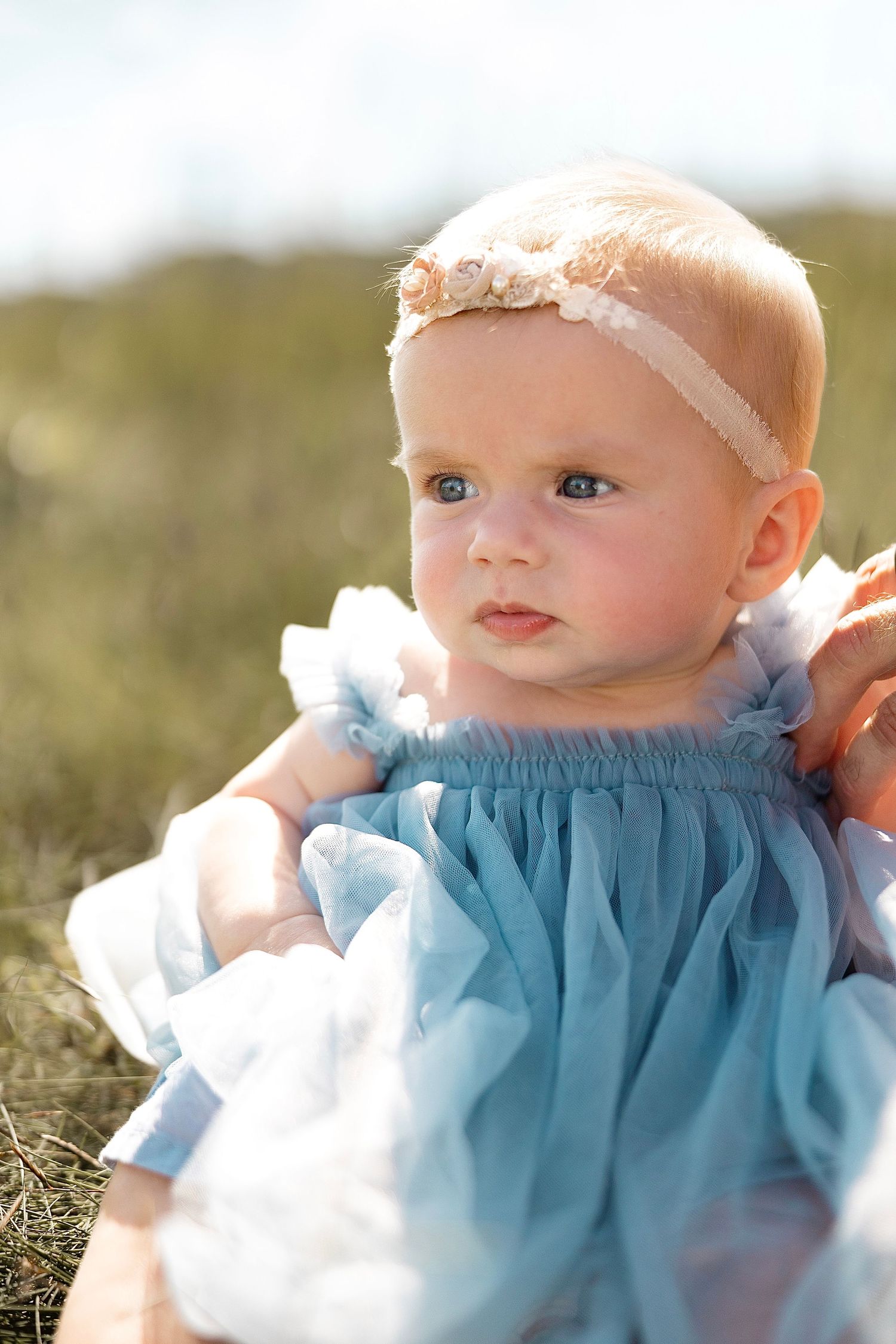 Adorable baby girl in blue tulle dress with floral headband outdoors in sunlight.