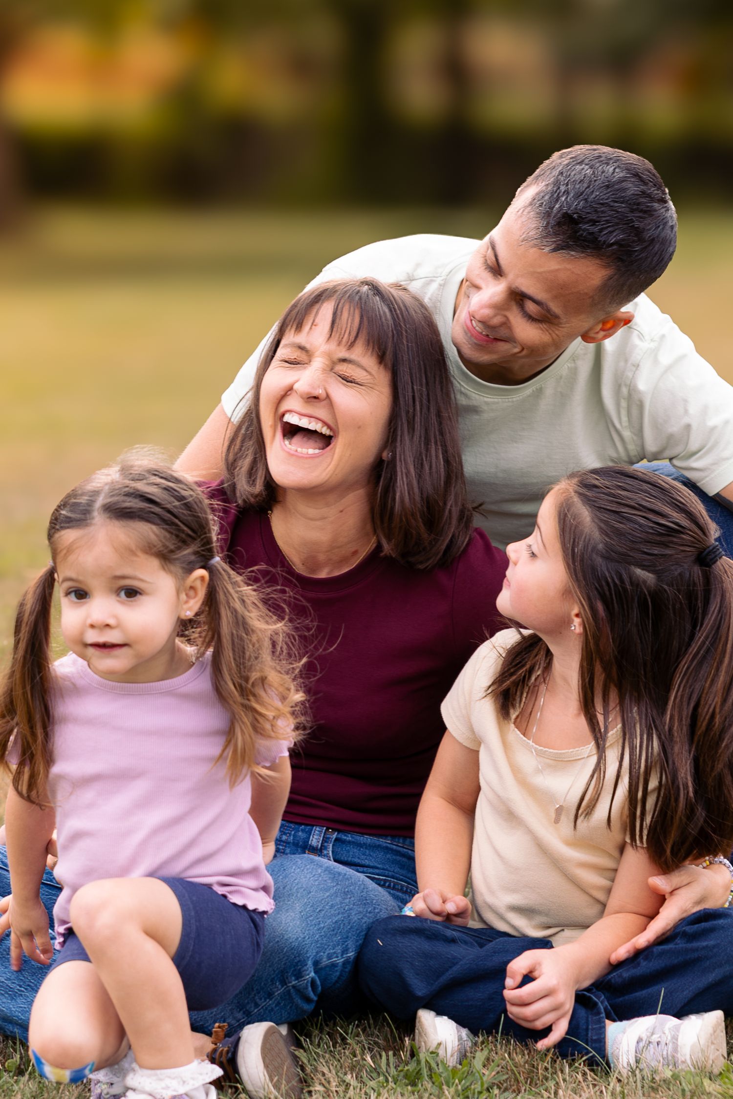 Happy family of four sitting on grass in a park, laughing and enjoying time together outdoors.